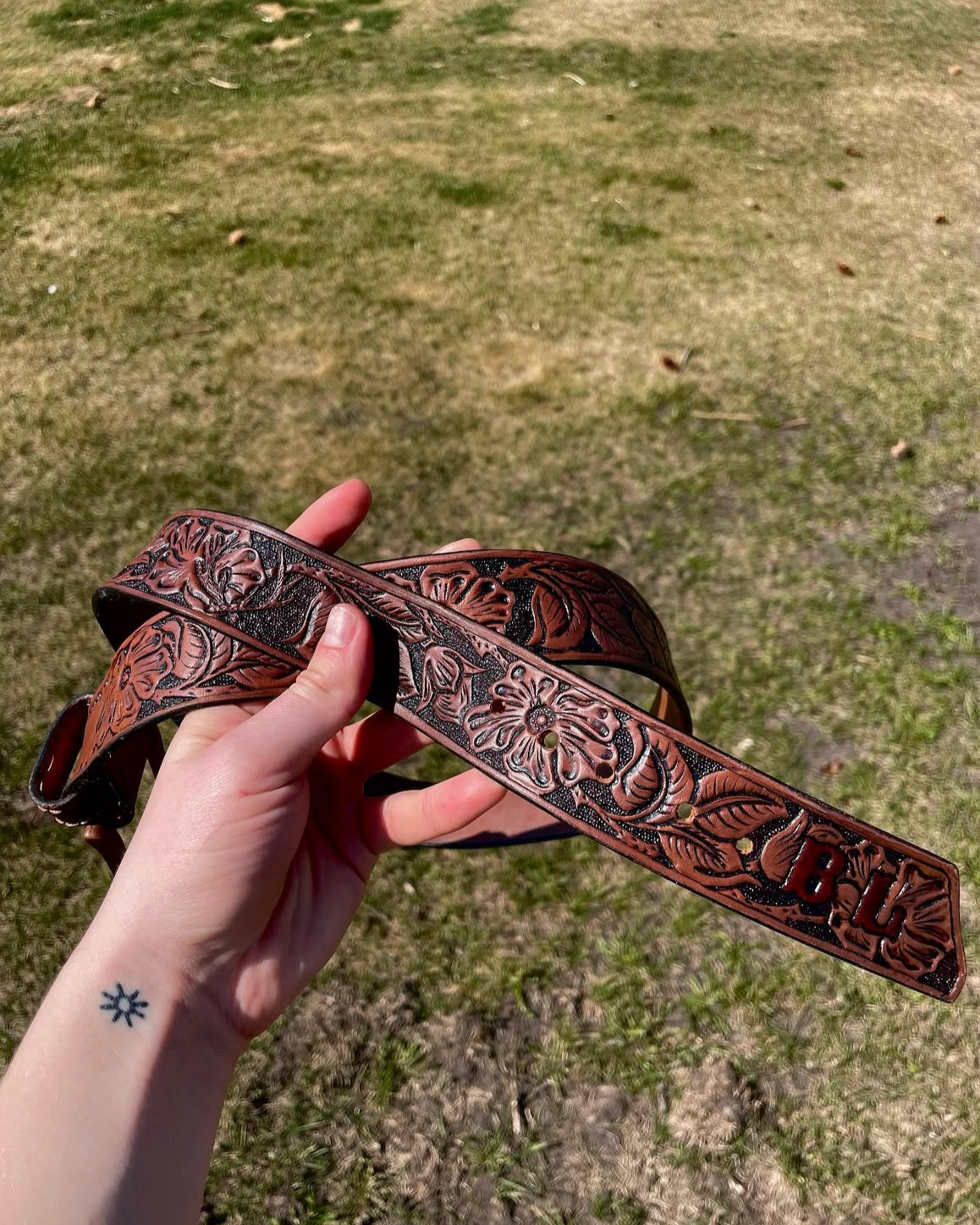 Hand holding a brown leather belt with floral embossing and the initials 'BL' engraved on it, outdoors on grass.