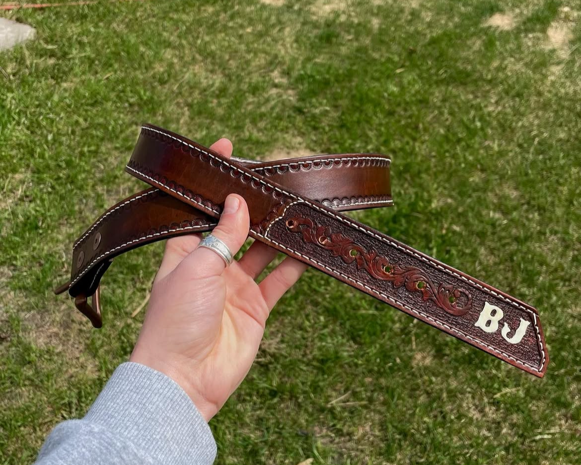 Hand holding a brown leather belt with an embossed design and the initials 'BJ' in metal detail, outdoors on grass.