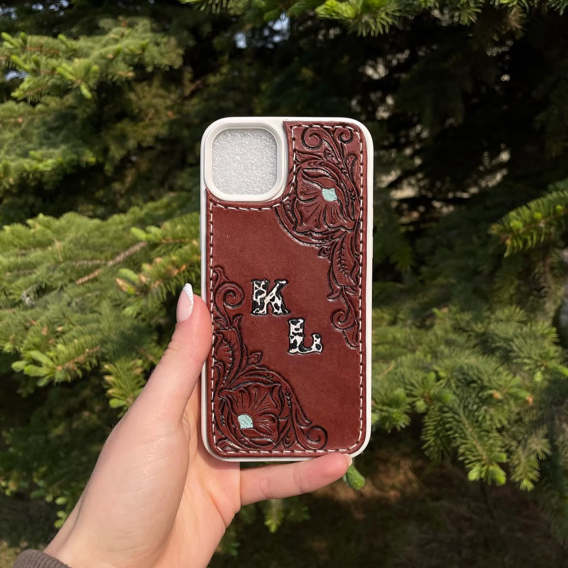A hand holds a smartphone with a brown leather case that has embossed floral designs and the initials L and K in a patterned font. The background features green pine tree branches.