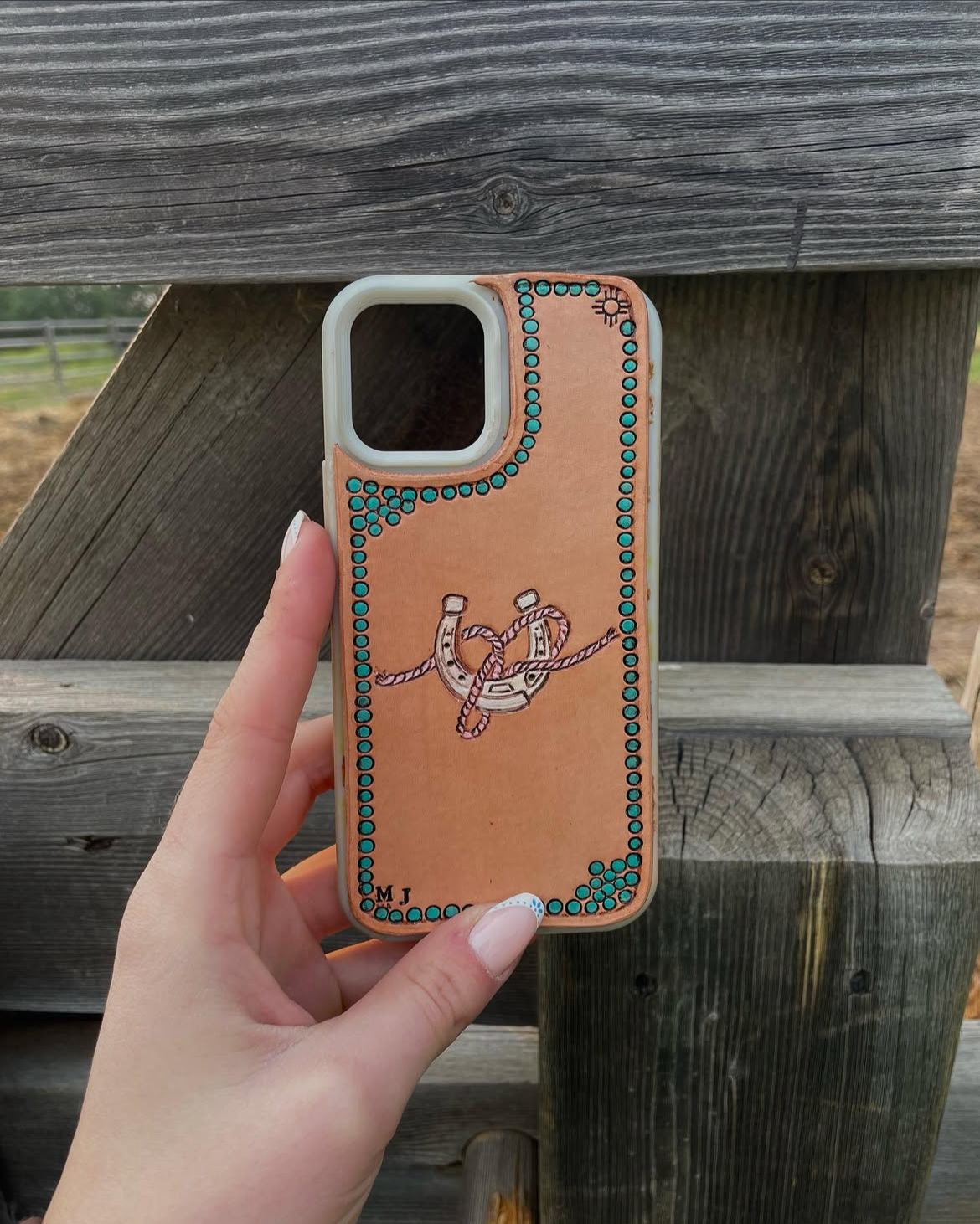A person holding an iPhone case with a rope and pulley design, featuring turquoise dots around the edges and initials 'M J' on the bottom left corner, against a wooden fence background.