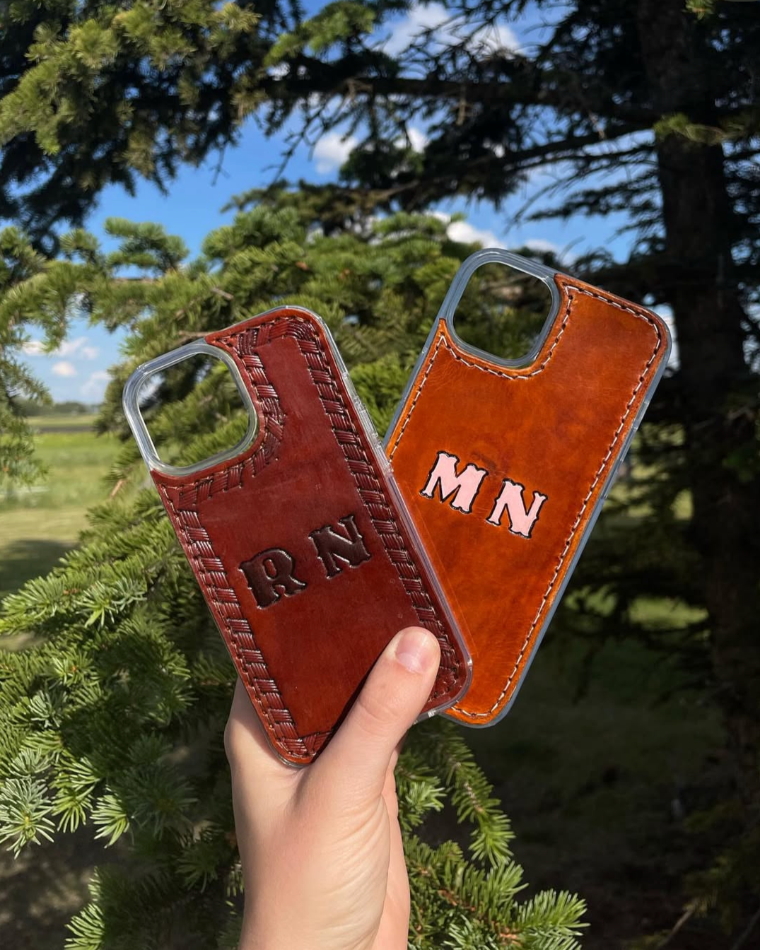 Two leather phone cases with stitched borders and embroidered initials, held against landscape with trees and blue sky in background.