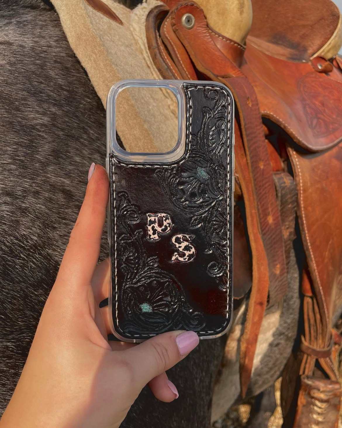 A hand holding a black engraved phone case with the letters 'P S' on it, behind a saddle and a horse's hide.