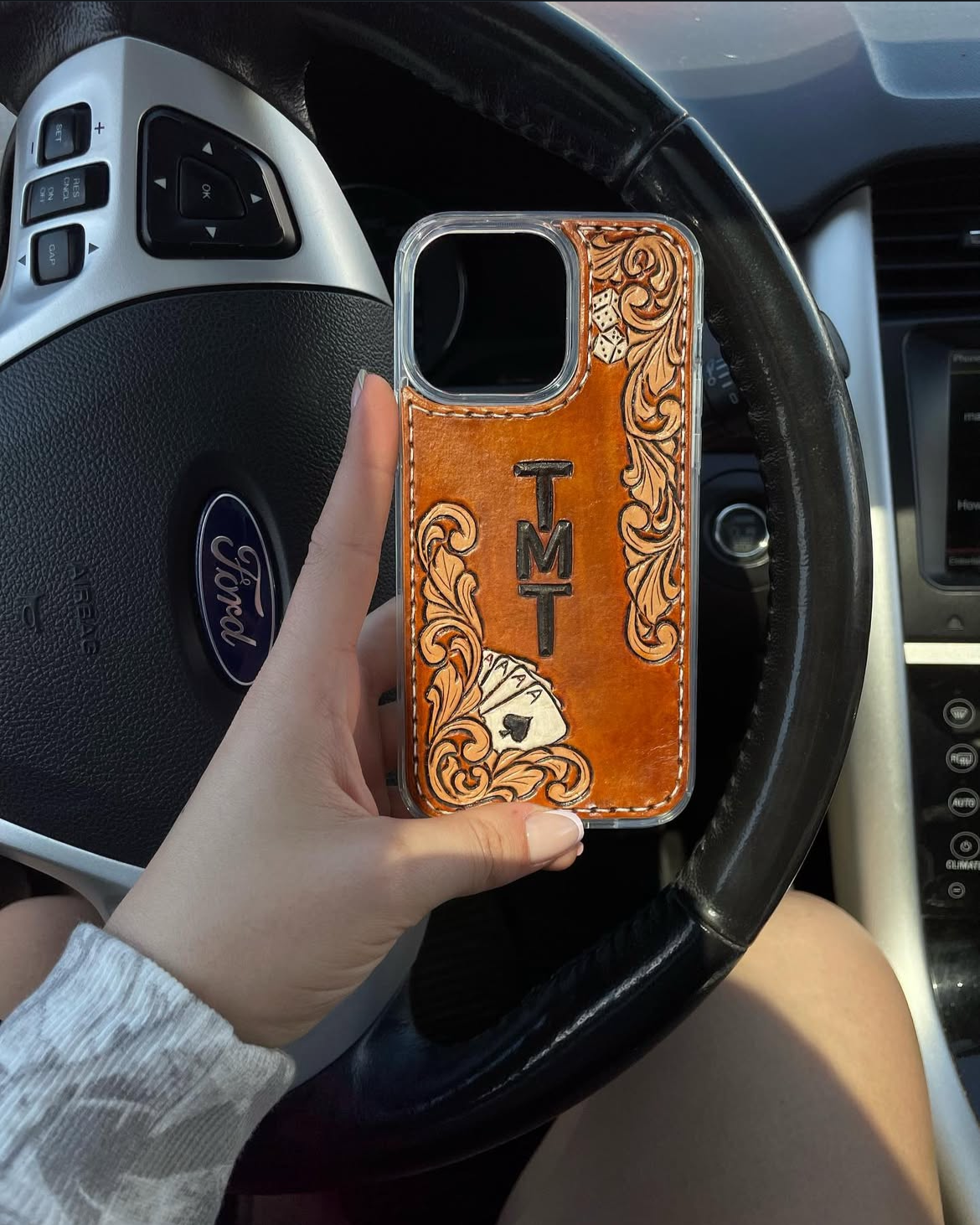 Hand holding a smartphone with a leather-textured case featuring the word 'TMT', playing cards, and decorative scrollwork inside a car at the steering wheel.