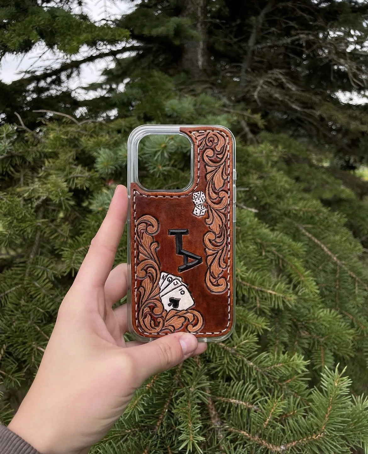 A hand holding a phone case with leather and wood designs, featuring poker symbols and a pair of dice, against a background of green pine branches.