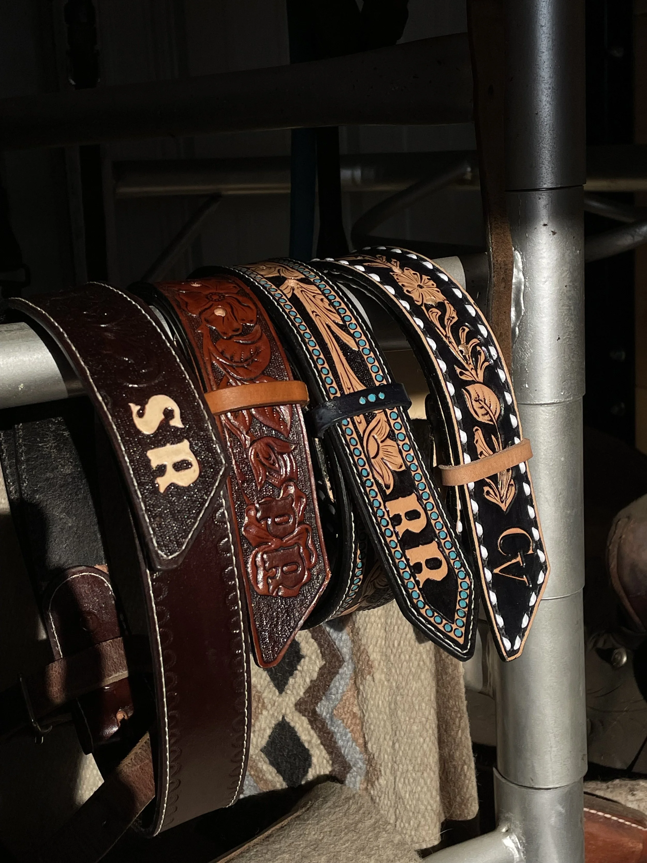 A display of five leather belts hanging on a metal rod with various patterns, colors, and embellishments.