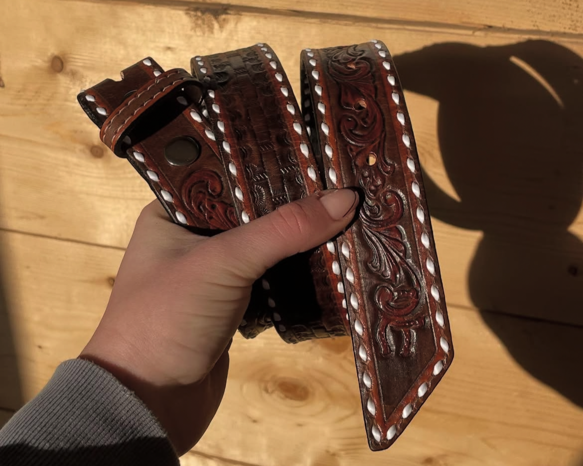 A hand holding a brown leather belt with intricate embossed designs and white stitching, against a wooden background.
