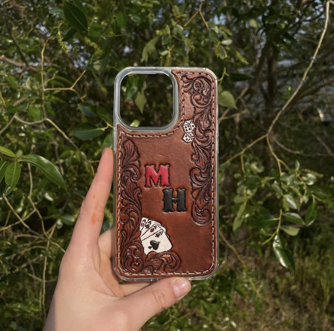 Handheld smartphone case with leather embellishments, embroidered initials 'M H', a pair of dice, and playing cards, against a background of green leafy trees.