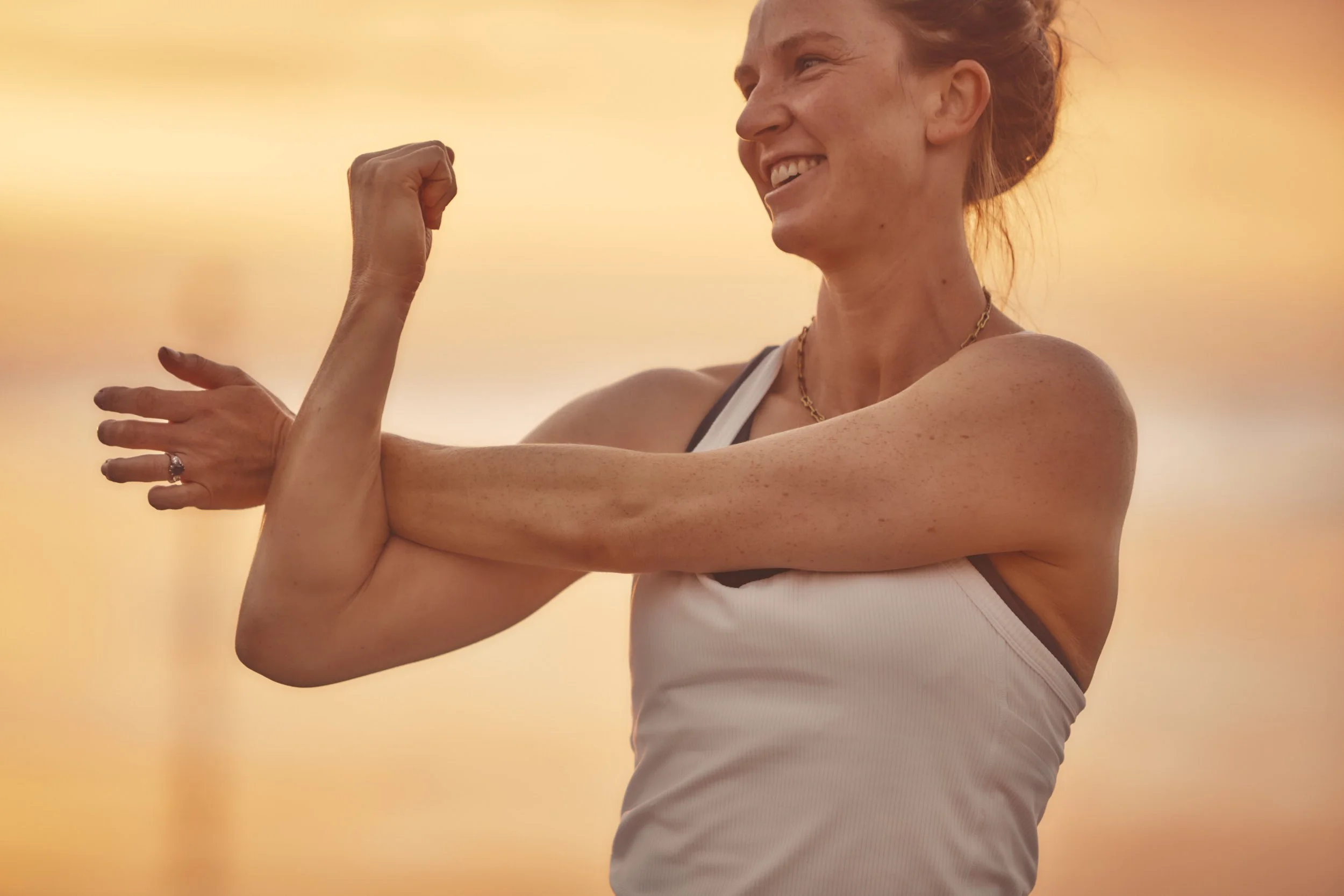 A woman with a joyful expression performing arm and shoulder exercises at sunrise.