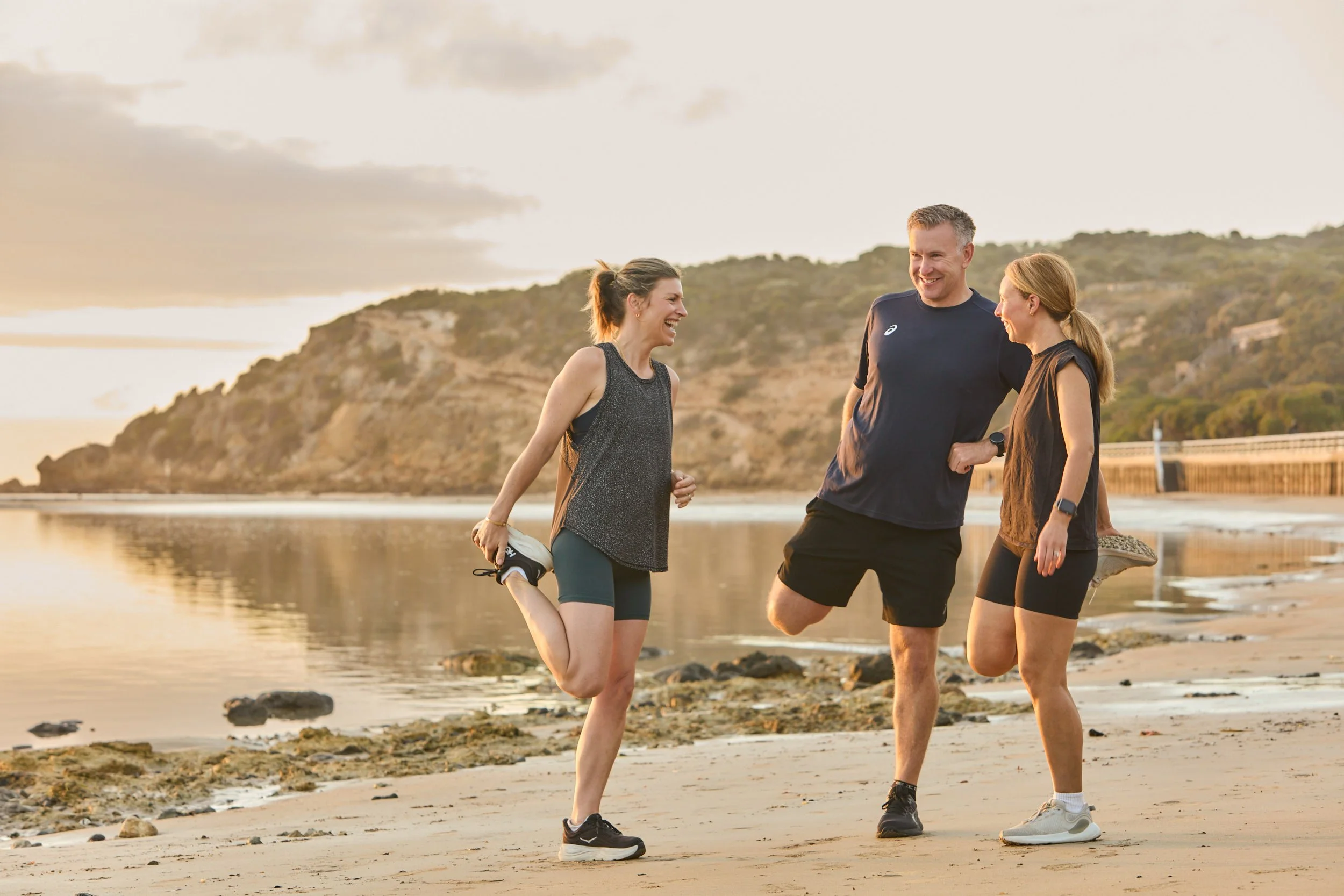 Three people, two women and one man, stretching on a beach at sunset, smiling and enjoying their workout.