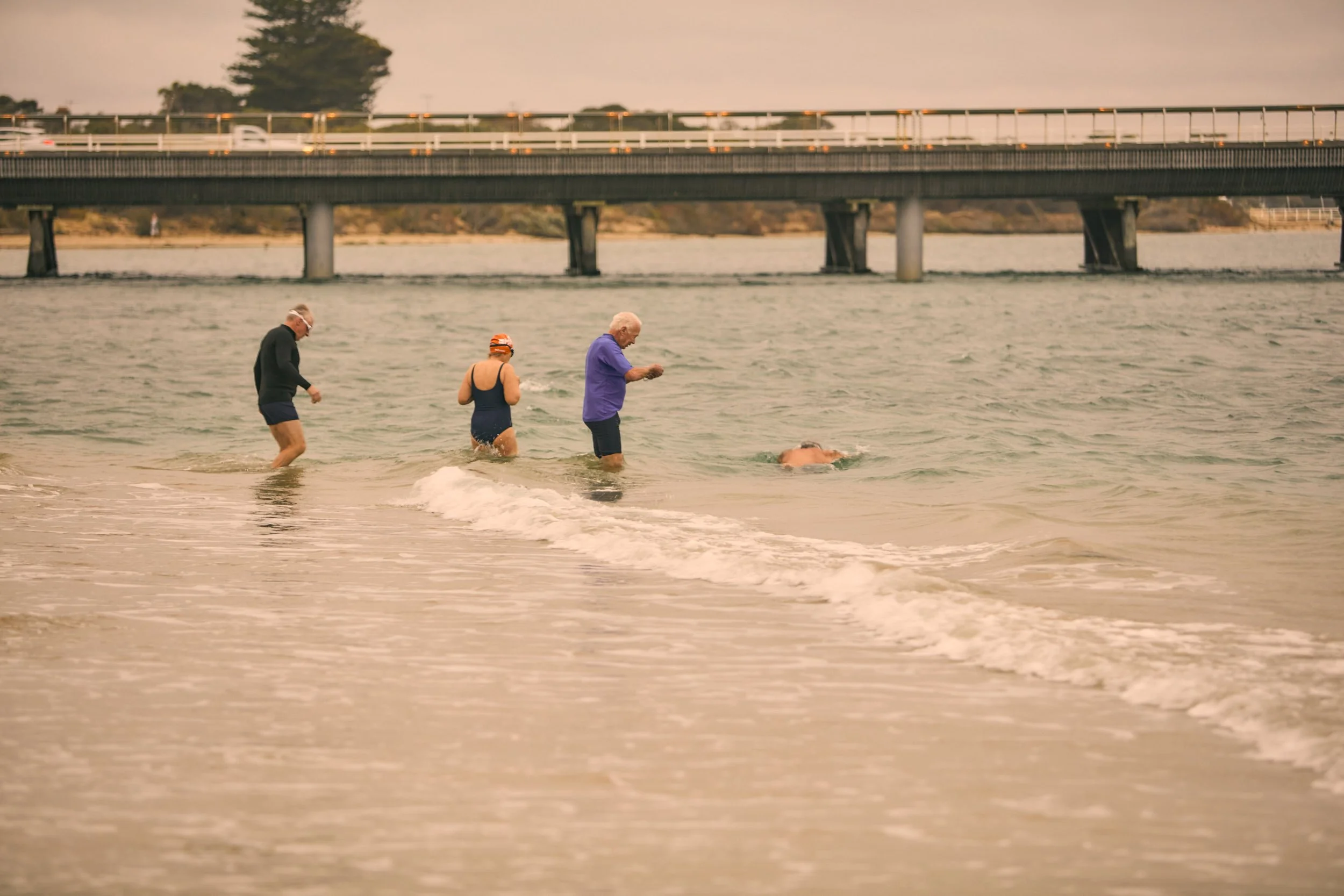 Four elderly people wading in the ocean near a sandy beach, with a Barwon Heads bridge in the background and an overcast sky.