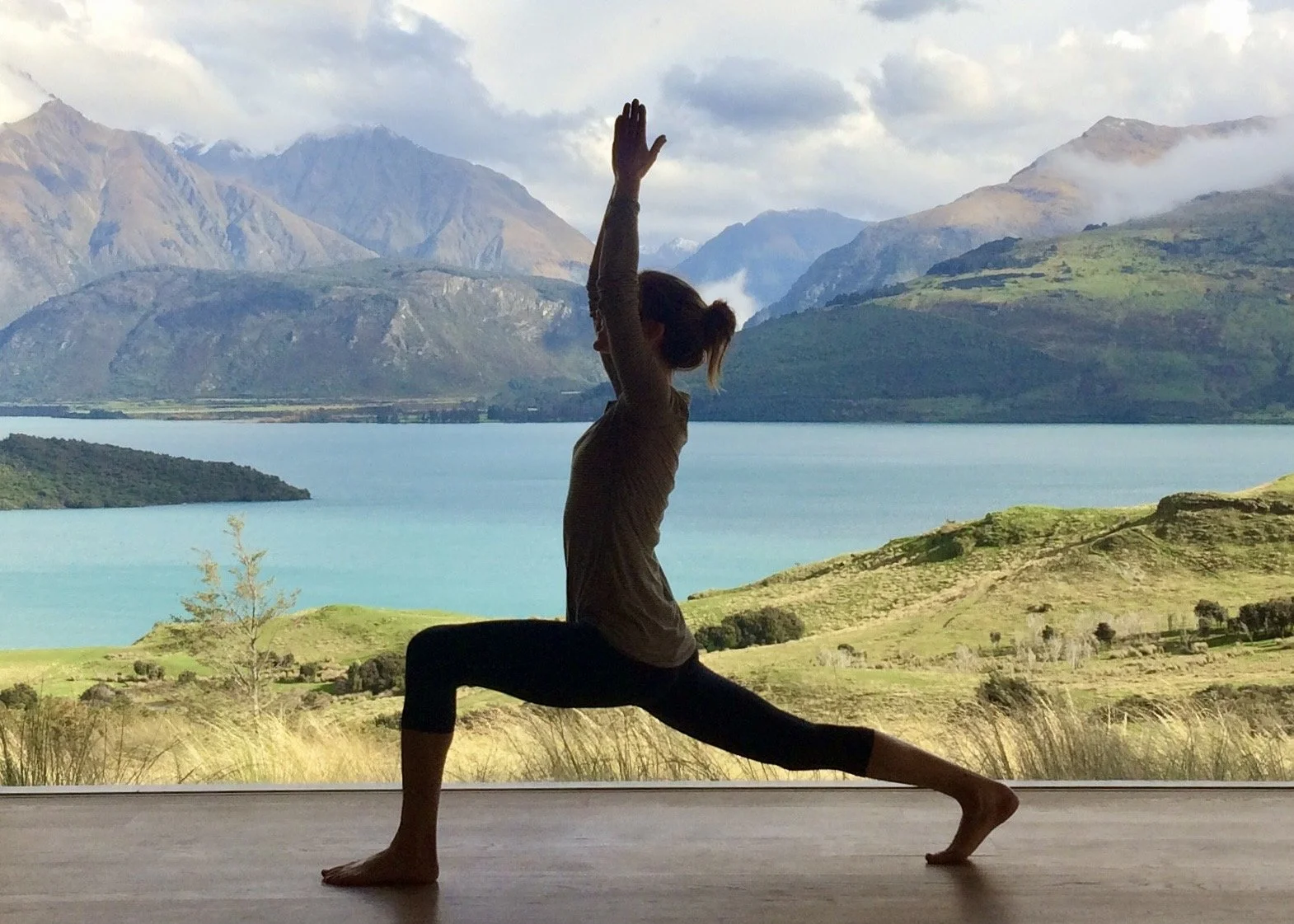 A woman practicing yoga indoors with a scenic mountain and lake view in the background.