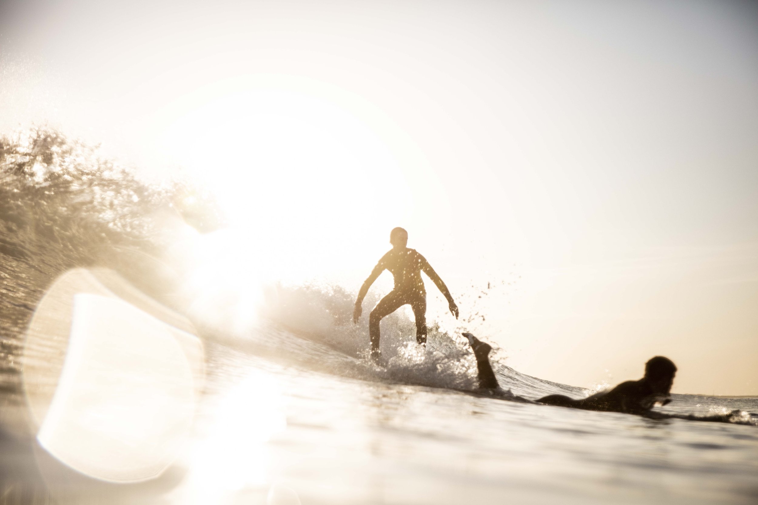 Silhouetted surfers riding a wave at sunset with bright sun in background