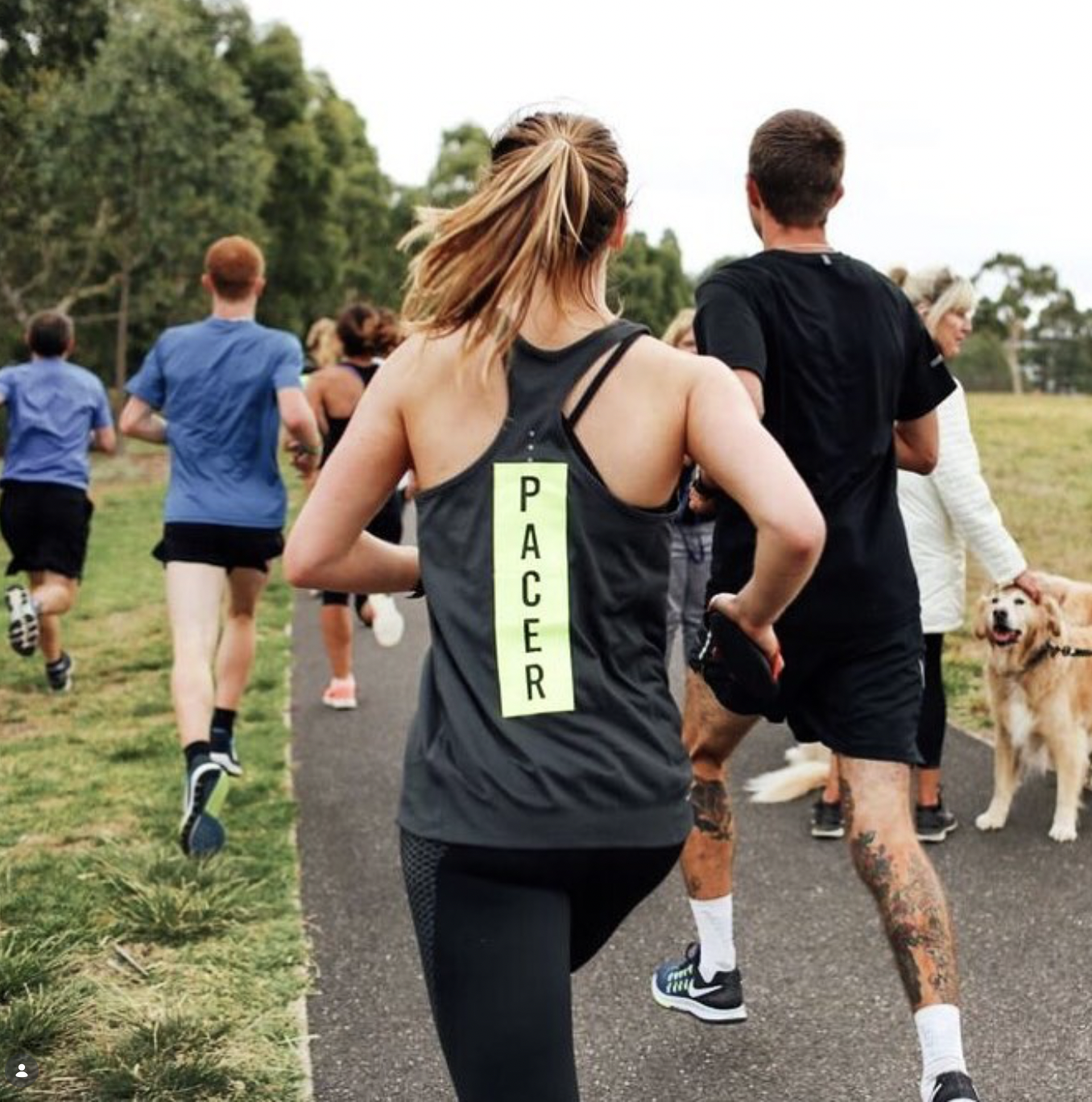 A group of people running outdoors on a trail with trees in the background, one woman in the foreground is wearing a black tank top with a yellow tag labeled 'PACER', some runners are wearing athletic clothes and shoes, and there are dogs walking with them.