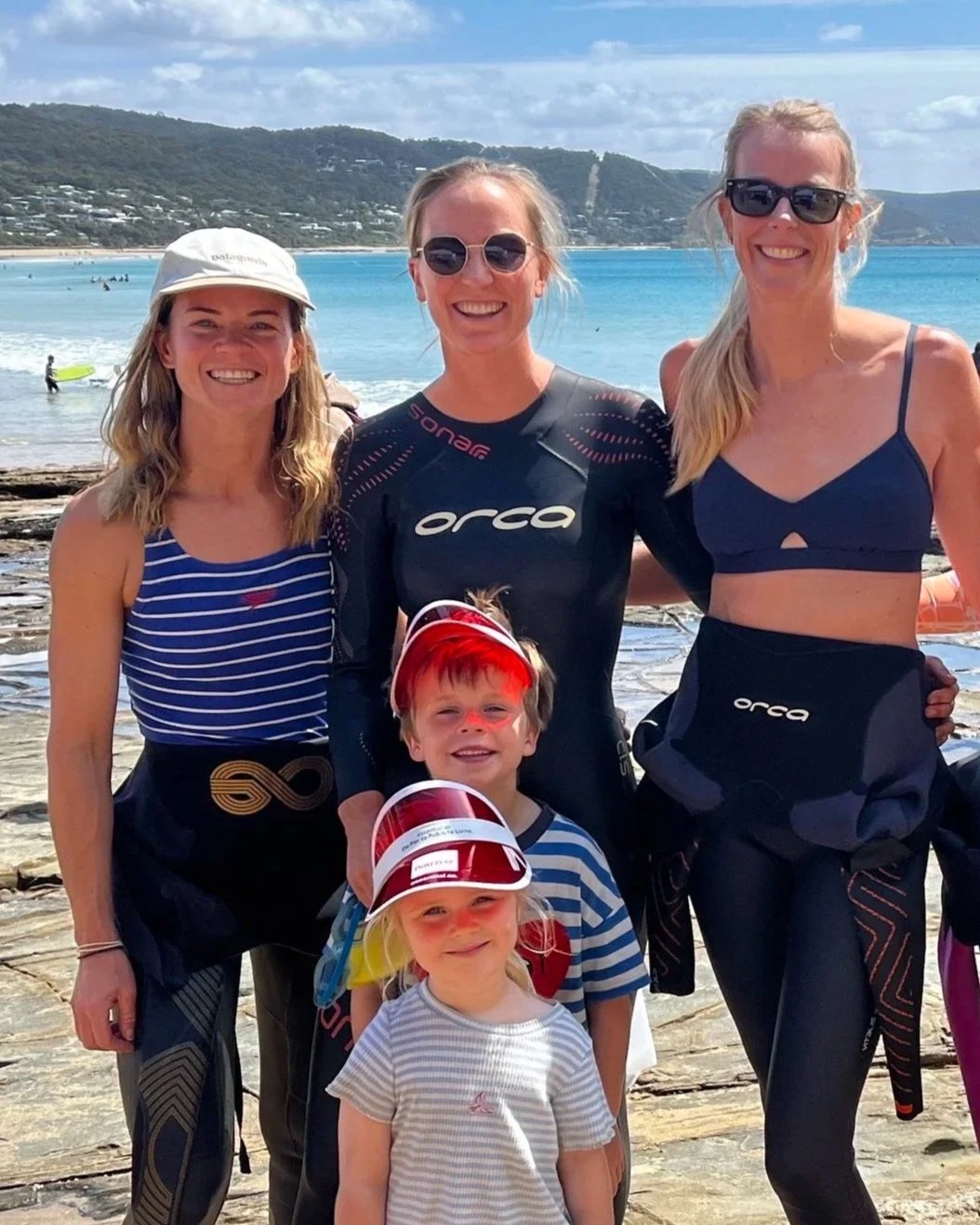 A group of five people, three women and two children, at the beach with the ocean, a line of hills, and beachgoers in the background. The women are dressed in swimwear and athletic gear, and the children are wearing striped shirts and hats. They are smiling and posing for the photo.