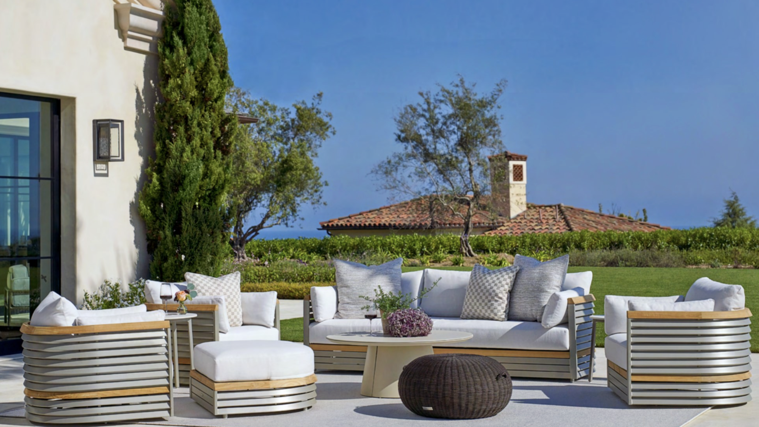 An outdoor patio setup with white cushioned furniture, pillows, and a round table with a flower arrangement, overlooking a garden with trees and a house with a tiled roof under a blue sky.