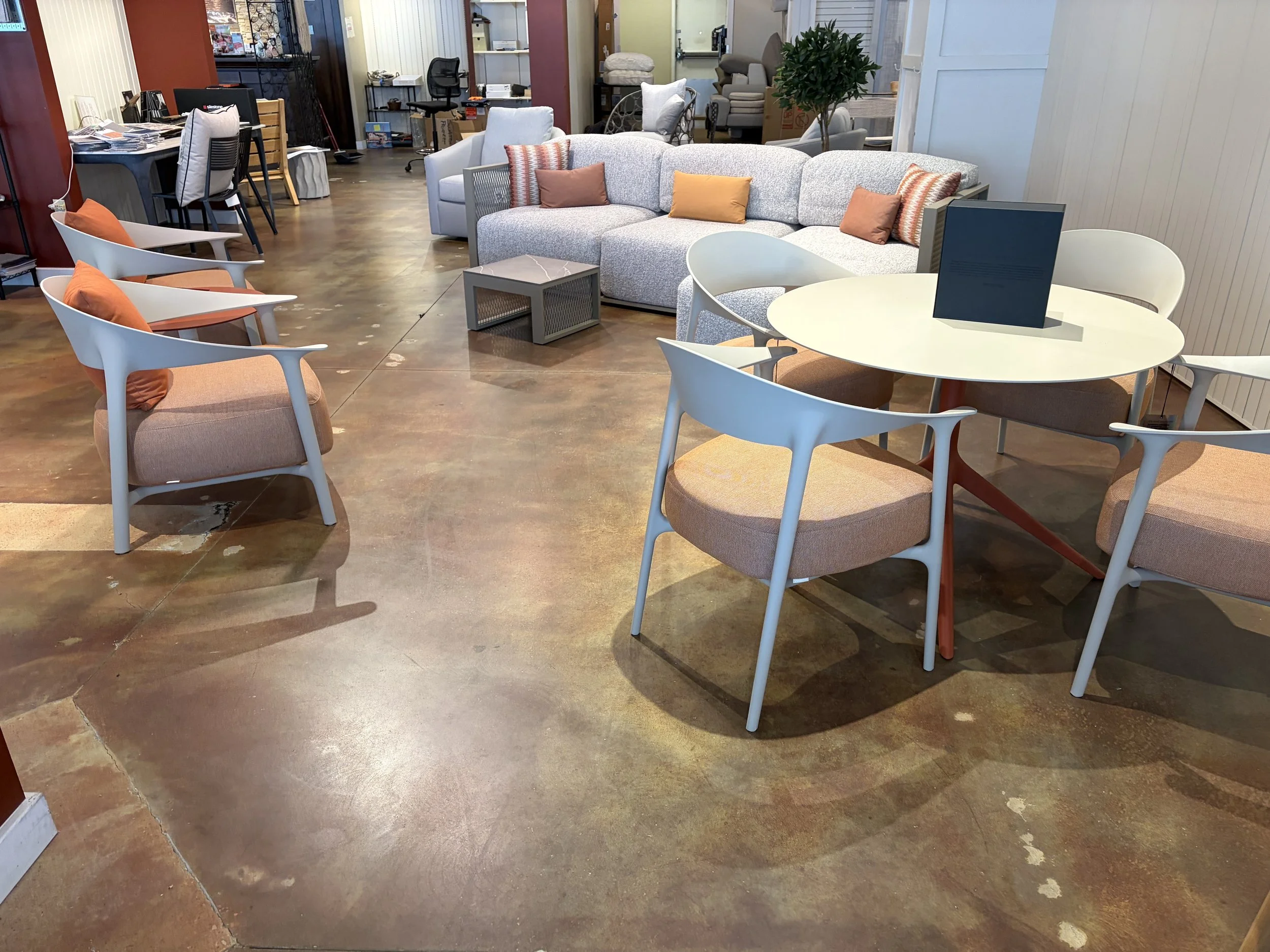 A living room with a white three-seater sofa, a round white dining table with chairs, and various other chairs and furniture in the background, with a polished concrete floor.