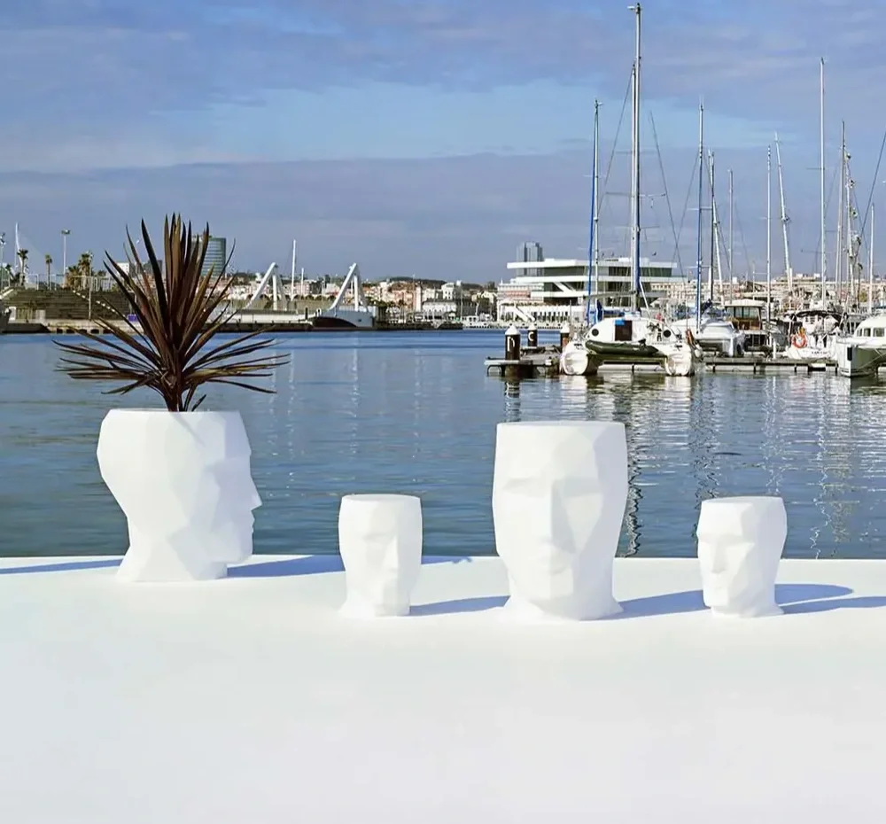 White artistic planters shaped like human heads with a plant in one of them, set on a white surface near a marina with boats and yachts in the background.