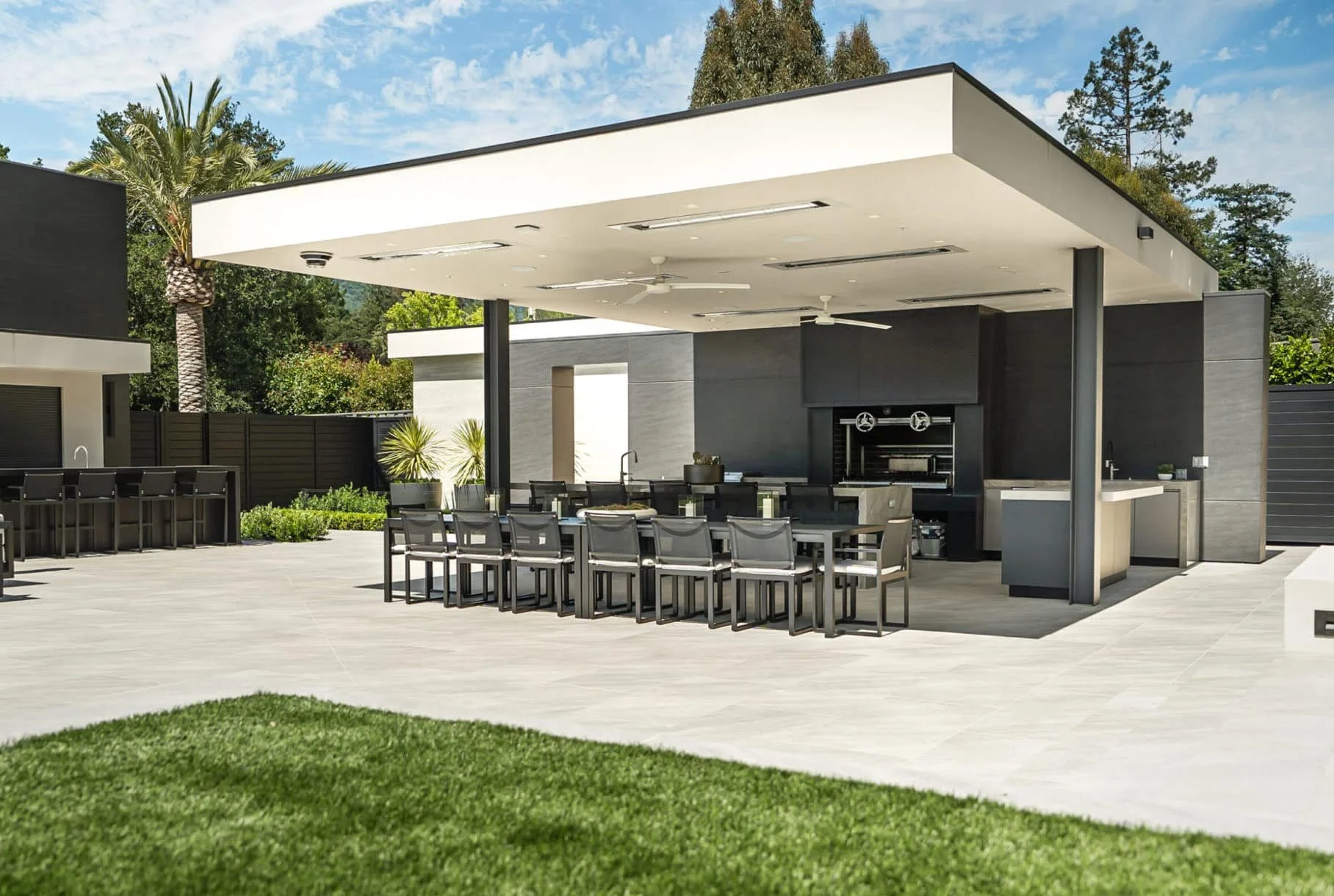 Modern outdoor patio area with a built-in grill and seating, surrounded by a sleek privacy fence and lush greenery, under a partly cloudy sky.