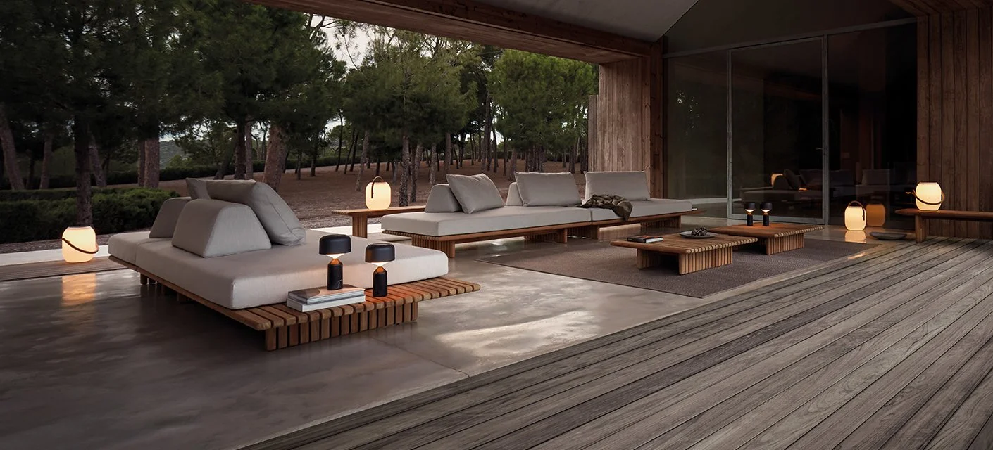 Modern outdoor patio with white cushioned seating, wooden tables, and lantern-style lights, overlooking a wooded area. Gloster