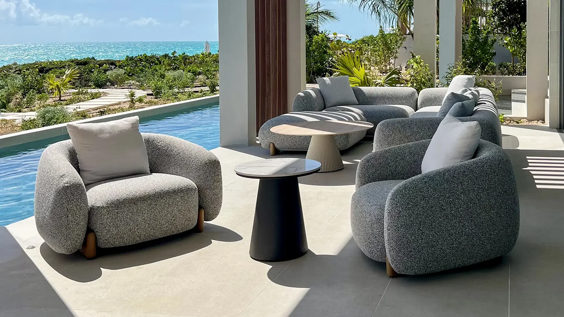 View of an outdoor seating area with modern grey armchairs with white pillows, and round black side tables, overlooking a swimming pool and lush green landscape with bushes and palm trees, with the ocean visible in the distance under a partly cloudy sky.