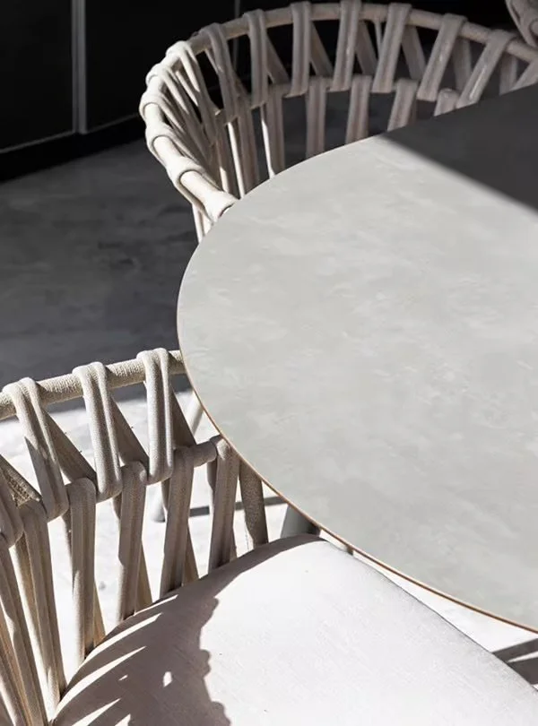 Part of a modern round dining table with a light-colored marble top and wicker chairs with beige cushions, indoors with natural light casting shadows.