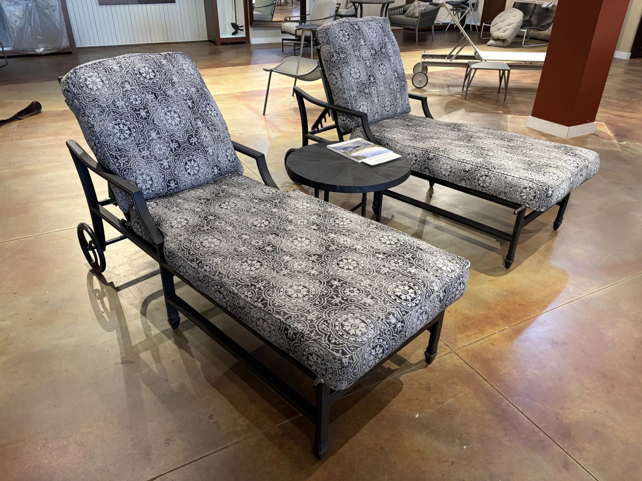 Two chairs with black metal frames and patterned fabric cushions set on a polished concrete floor in a showroom or furniture store, with a small round black table holding magazines between them, and various other furniture pieces visible in the background.