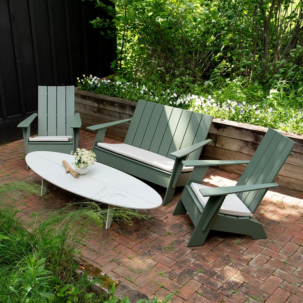 A cozy outdoor patio with green Adirondack chairs and a white oval table, decorated with flowers, on a red brick surface bordered by lush greenery and a flower bed.