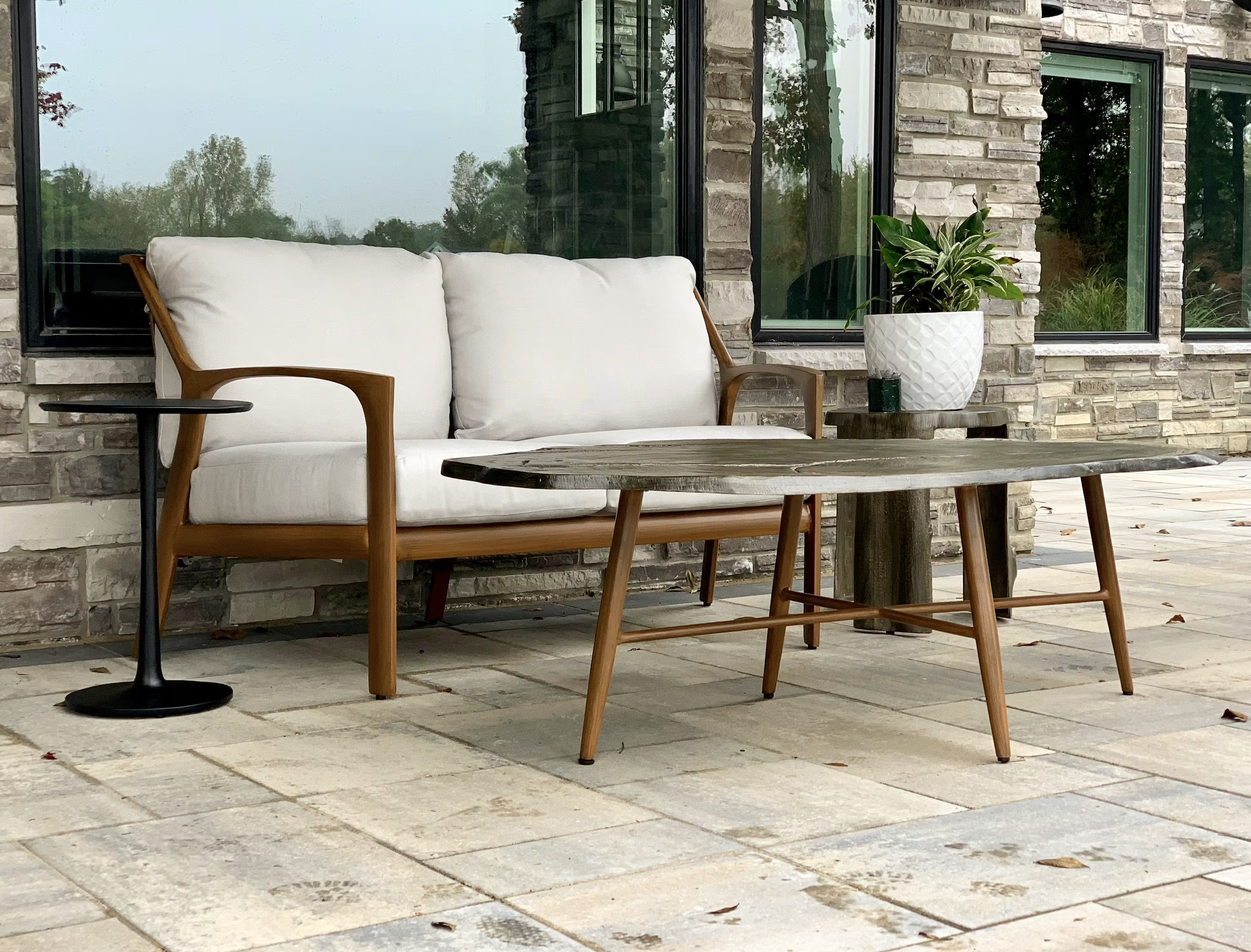 Outdoor patio area with a white cushioned outdoor sofa, a black round side table, a wooden coffee table, and a large potted plant in front of a stone house with large windows reflecting trees.