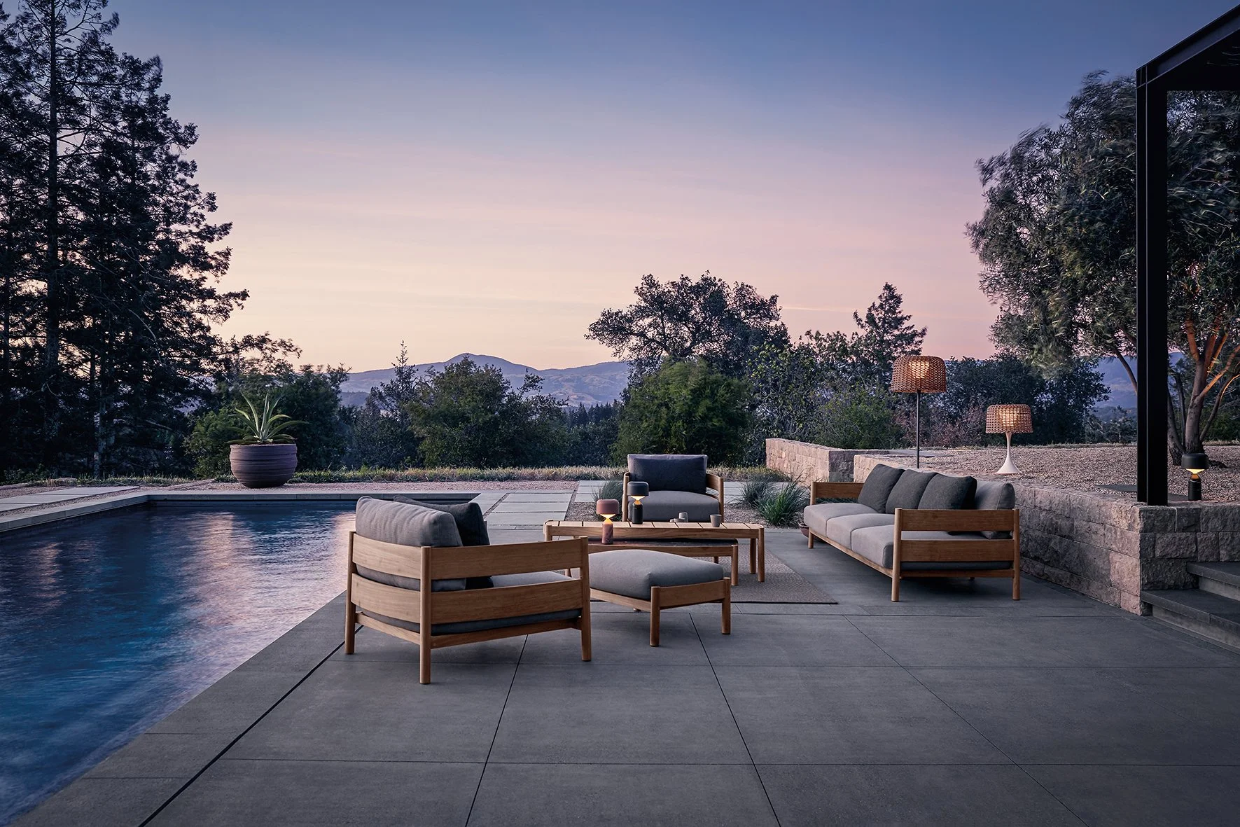 A modern outdoor patio with a swimming pool, outdoor seating including a sofa and chairs with cushions, two table lamps with woven shades, potted plant, and lush trees with mountains in the background at dusk. Gloster