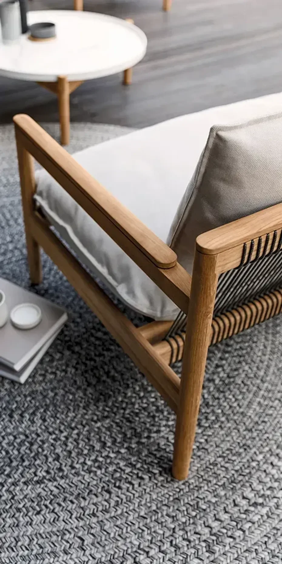 Wooden armchair with light gray cushion and beige pillow on a gray woven rug, next to a small tray table with candles and books, in a modern living room.