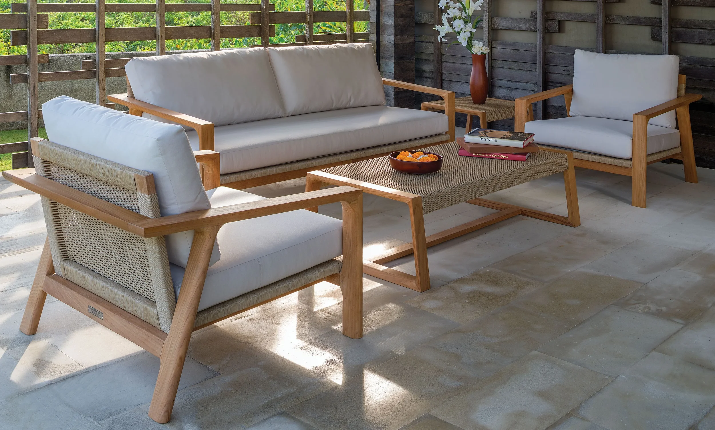 An outdoor patio setup with a white cushioned sofa, a matching armchair, and a wooden armchair with white cushions. There is a rectangular woven coffee table with a bowl of decorative flowers and a couple of books on top. A wooden side table with a vase of white flowers is also present. The patio is enclosed with a wooden lattice and has a green view beyond.
