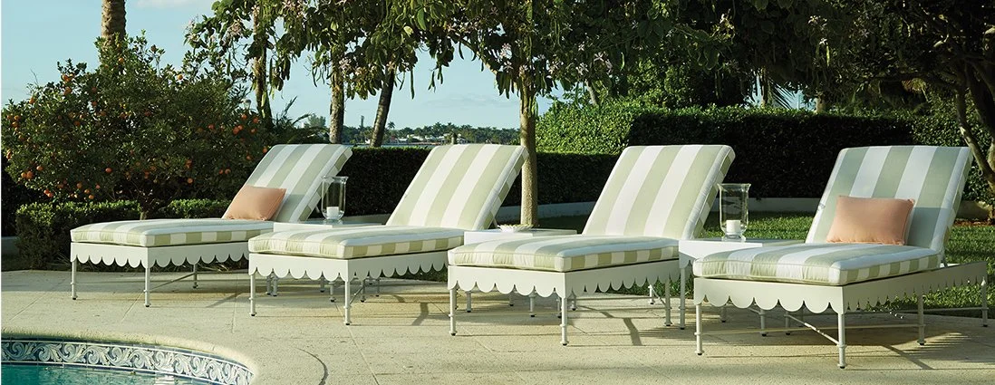 Four striped poolside lounge chairs with pillows and glasses of water on small side tables, arranged outdoors near a swimming pool, with greenery and trees in the background.
