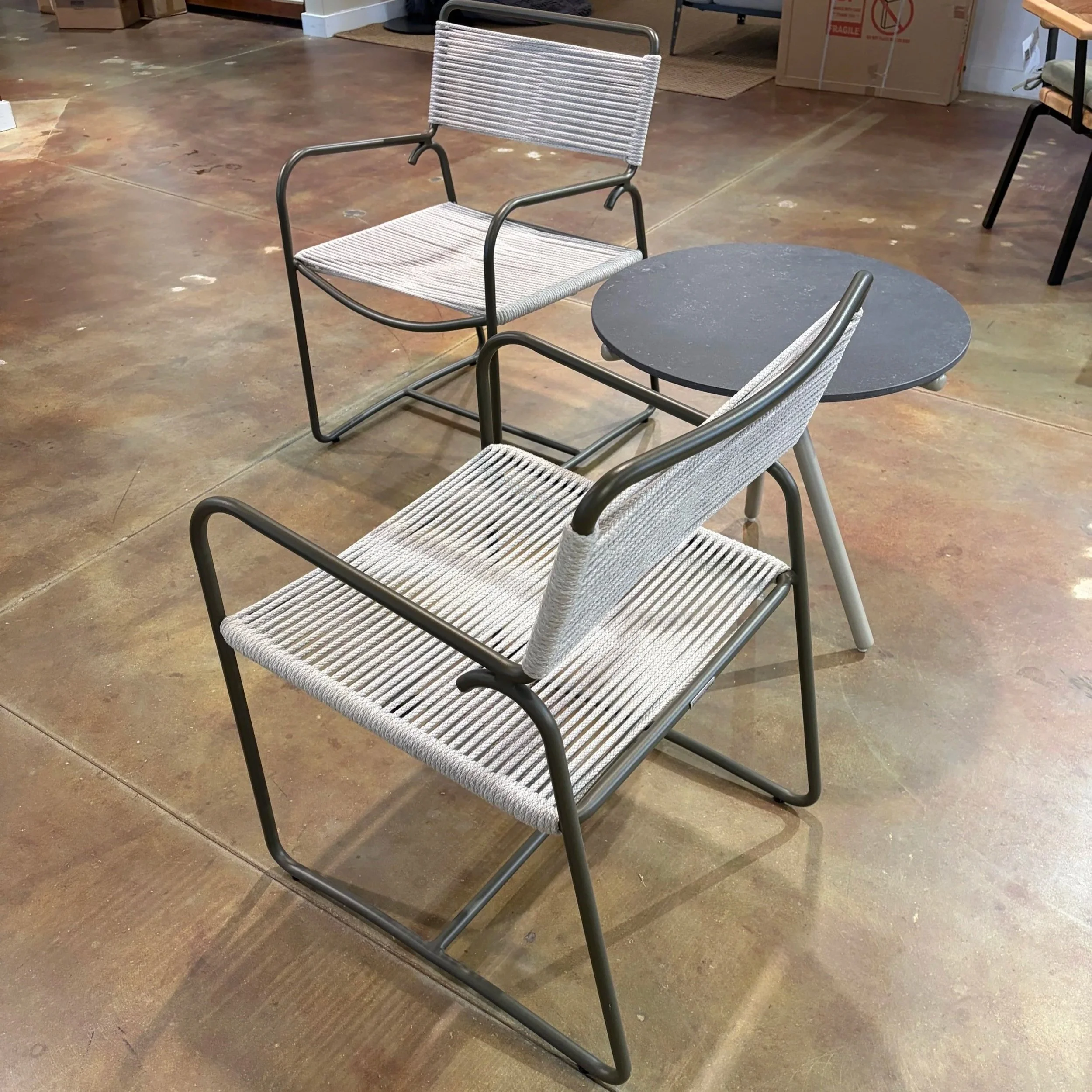 A metal and woven chair with armrests and a black round table beside it, situated on a concrete floor.
