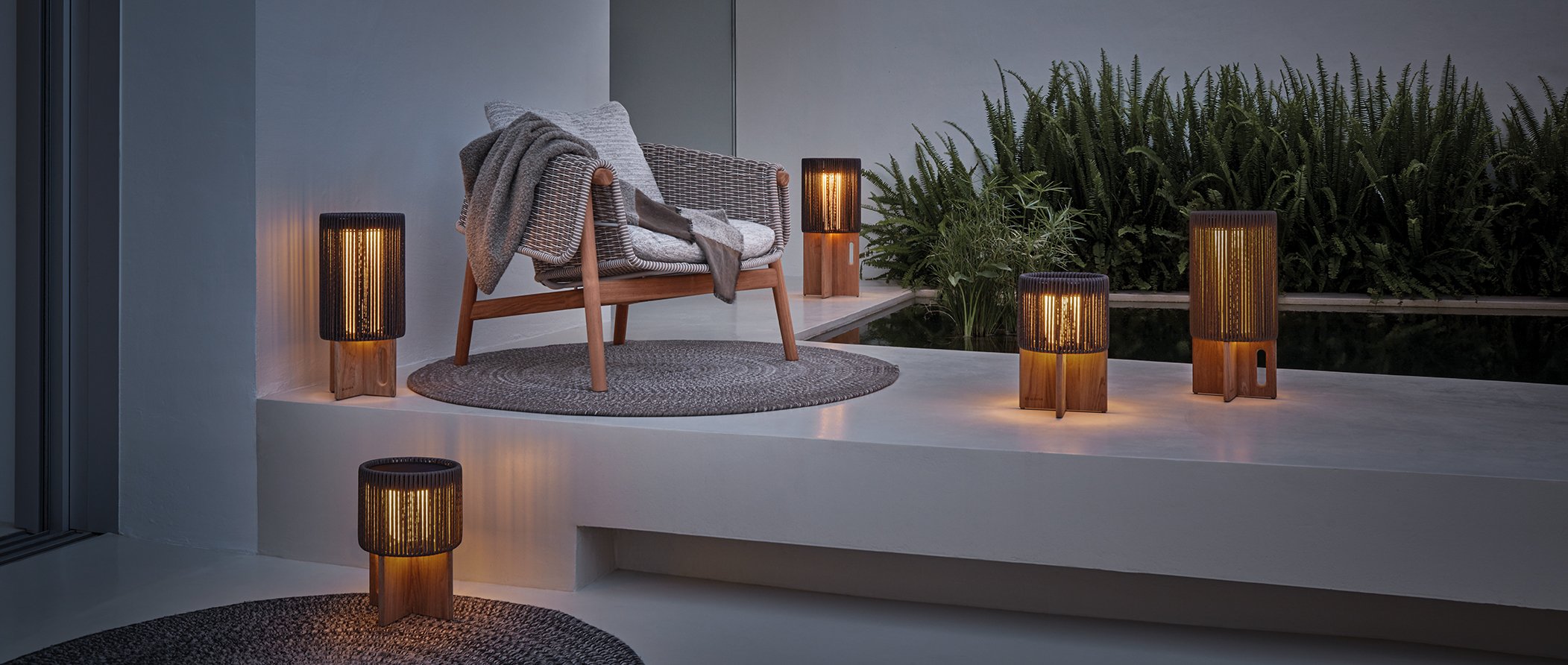 A cozy outdoor patio with a wooden bench draped with a gray blanket and pillows, surrounded by warm lighting from several lamps and lanterns, and lush green plants in the background.