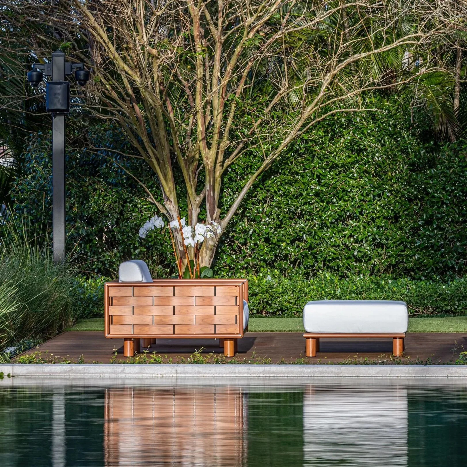 A modern outdoor scene featuring a wooden deck next to a body of water with two white chairs, a wooden storage box, a large tree with partly bare branches, lush green bushes and trees in the background, and a security camera on a pole.