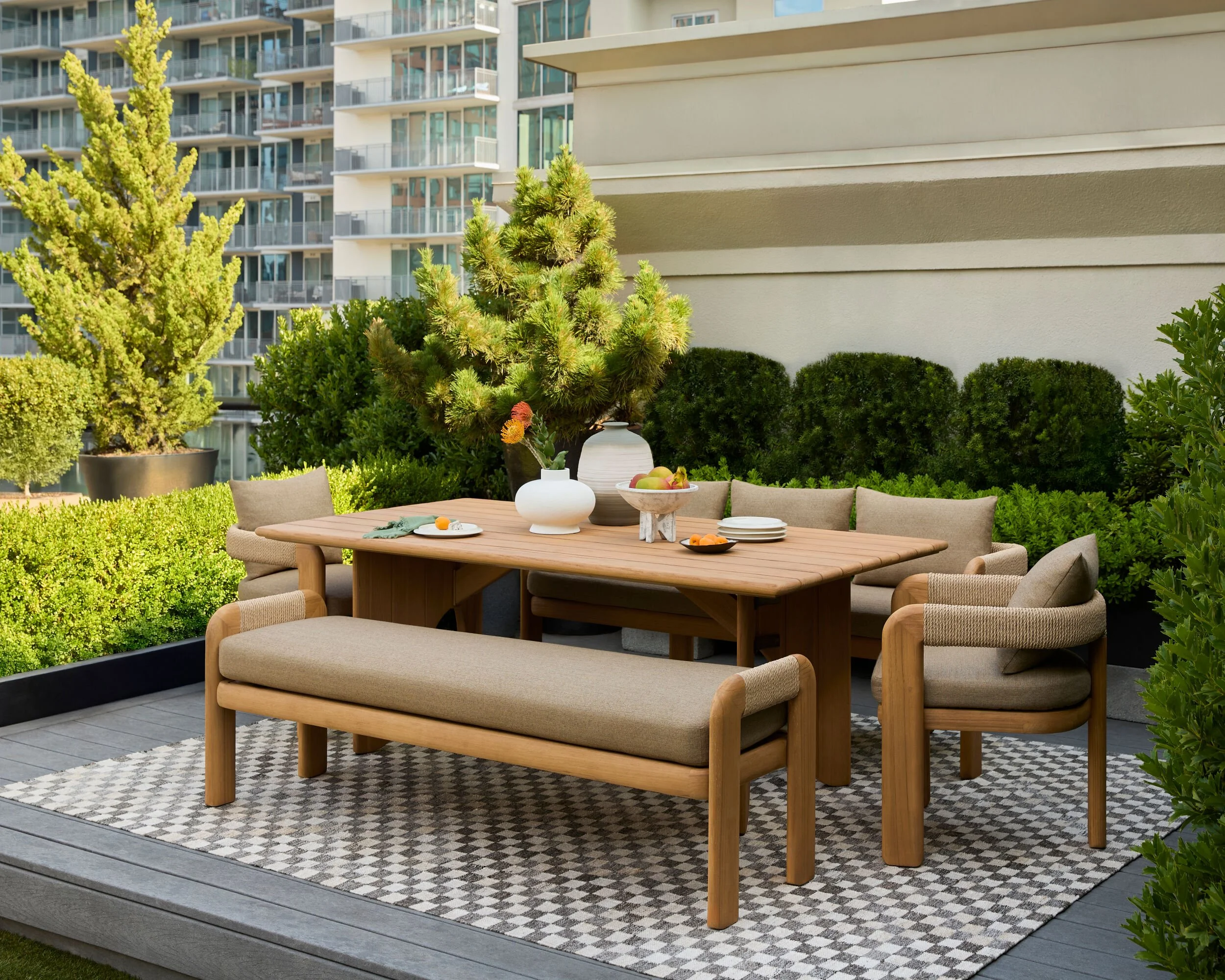 Outdoor balcony space with wooden dining table, beige chairs, potted plants, and apples, surrounded by green bushes and trees, with a modern apartment building in the background.