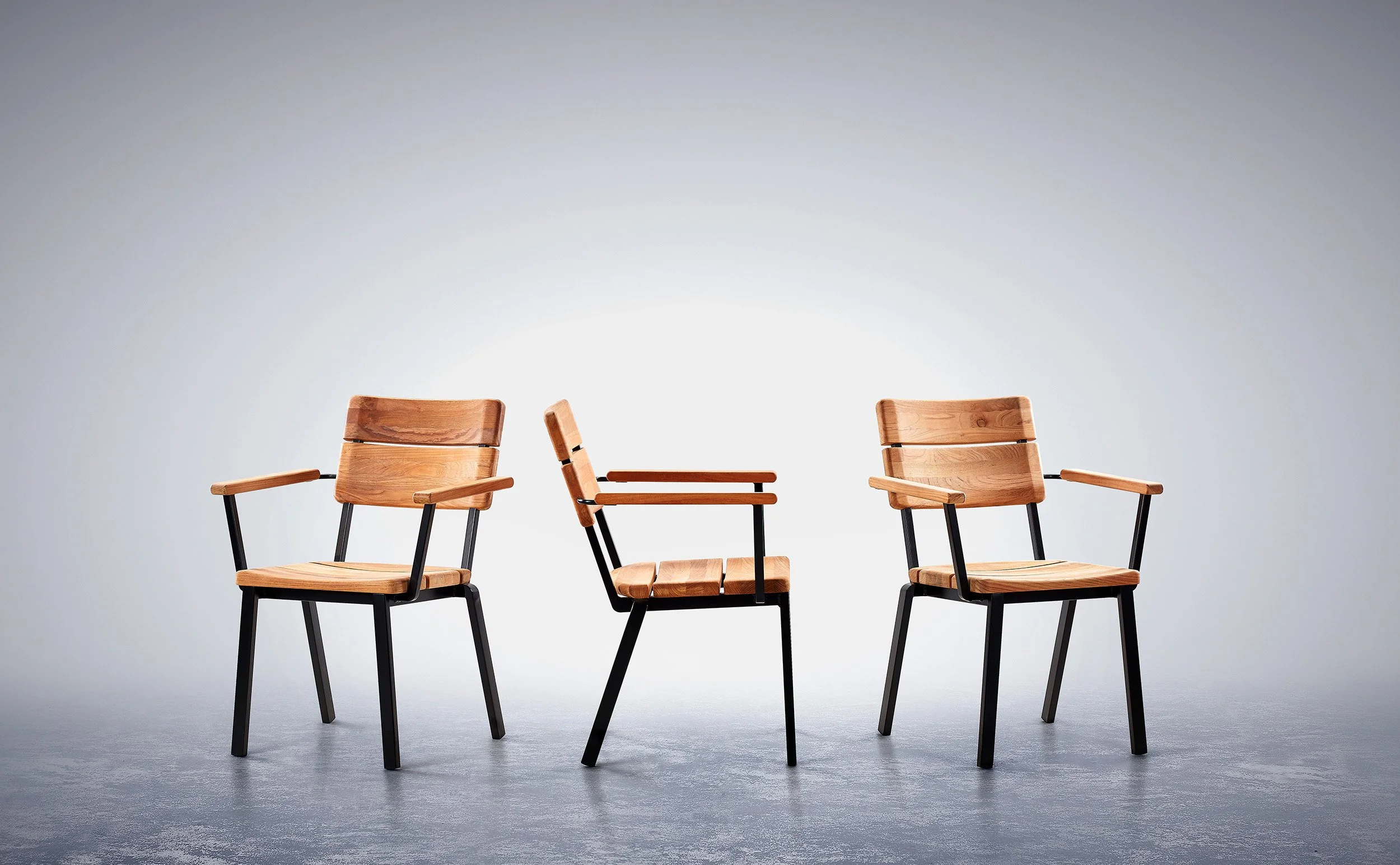 Four wooden chairs with black metal frames are arranged with three on the right side and one on the left side of the image, set against a plain light gray background. Barlow Tyrie