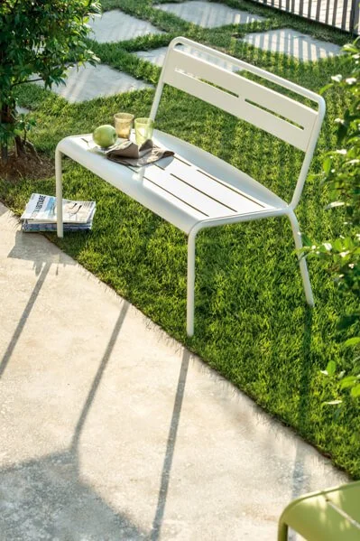 White outdoor bench with a tray holding a green apple, glass, and a cup on it, situated on a grassy patch next to a concrete walkway, with a book on the ground nearby.
