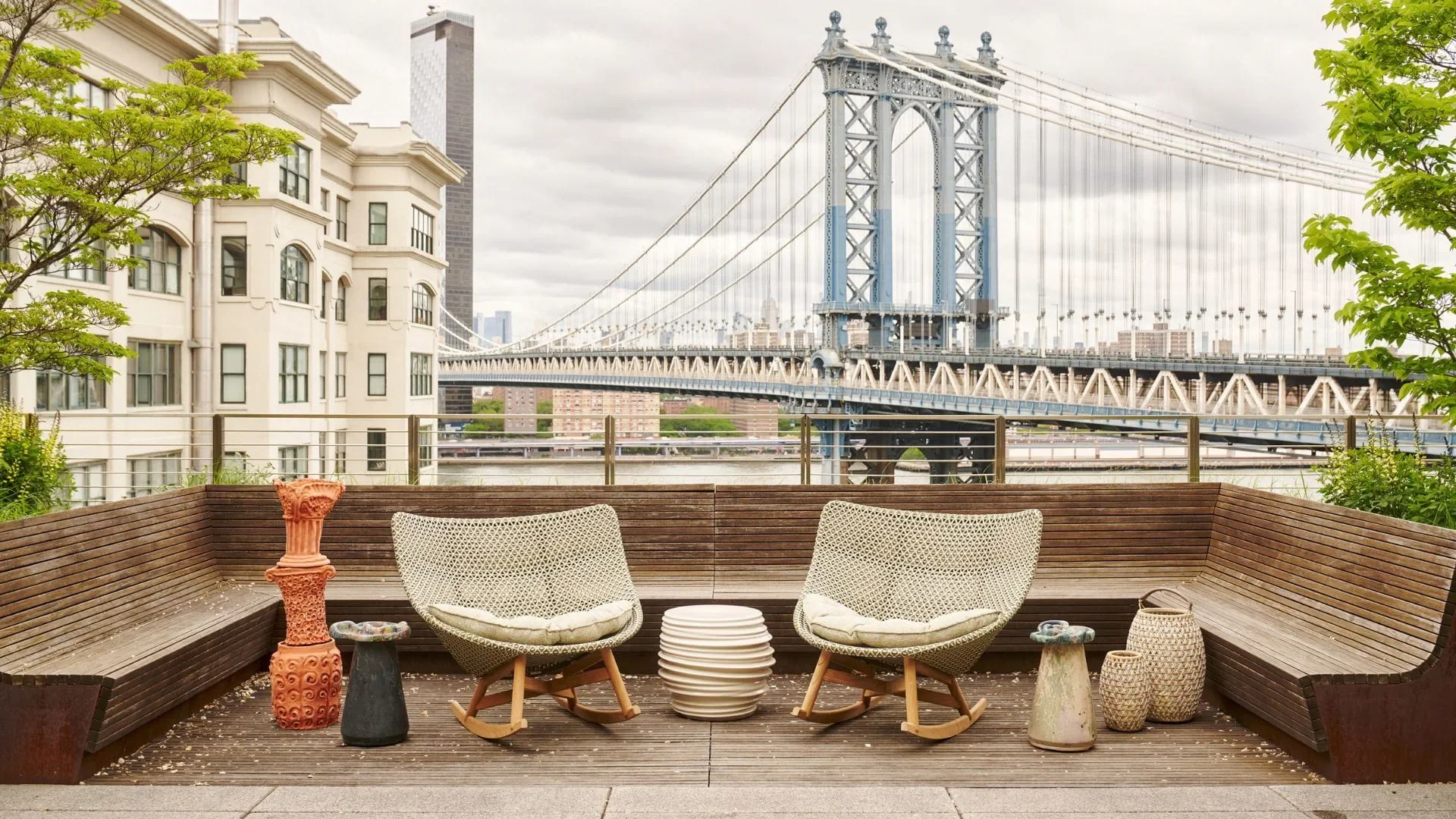 A rooftop deck with two wicker chairs, a low circular side table, and decorative vases. In the background, the Manhattan Bridge and city skyline of New York City are visible under a cloudy sky, with some greenery framing the scene.