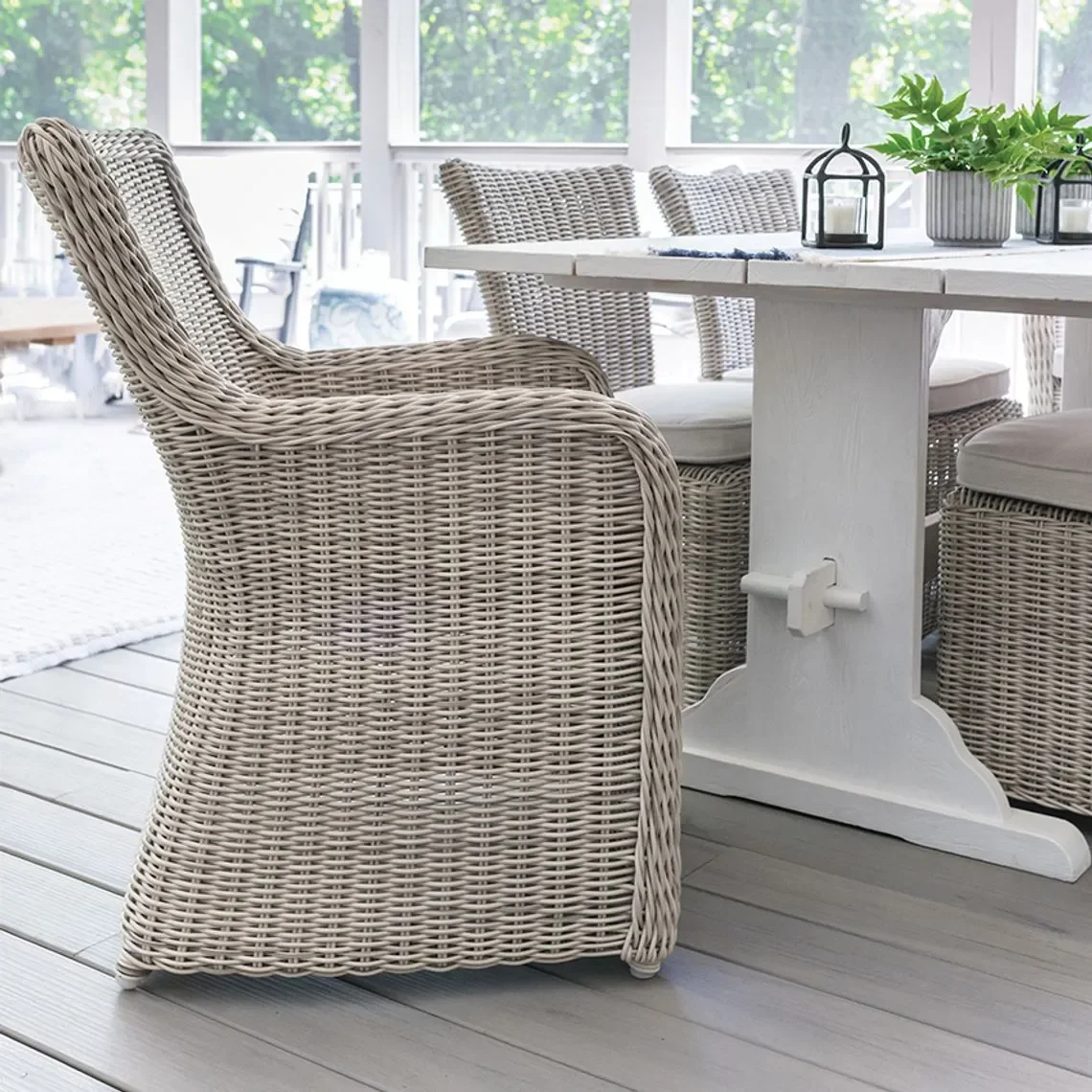 Br wicker chairs around a white table with potted plants and small lanterns on a porch or sunroom with large windows and greenery outside.