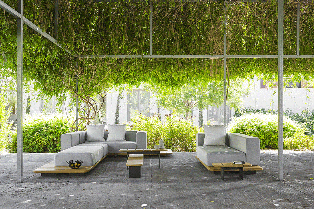 Outdoor seating area with two gray sofas, wooden coffee table, and lush green trees and bushes in the background.