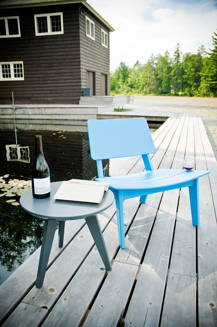 Blue chair and gray side table with a bottle of wine, an open book, and a glass of red wine on a wooden dock by a pond, with a dark brown house and green trees in the background.