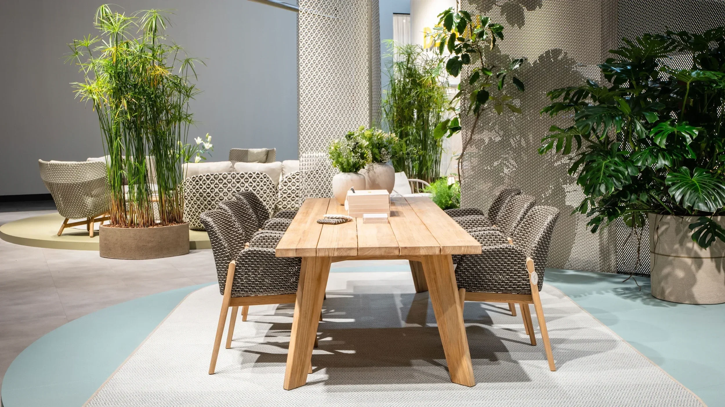Indoor space with a large wooden table surrounded by woven chairs, various green potted plants, and decorative privacy screens.