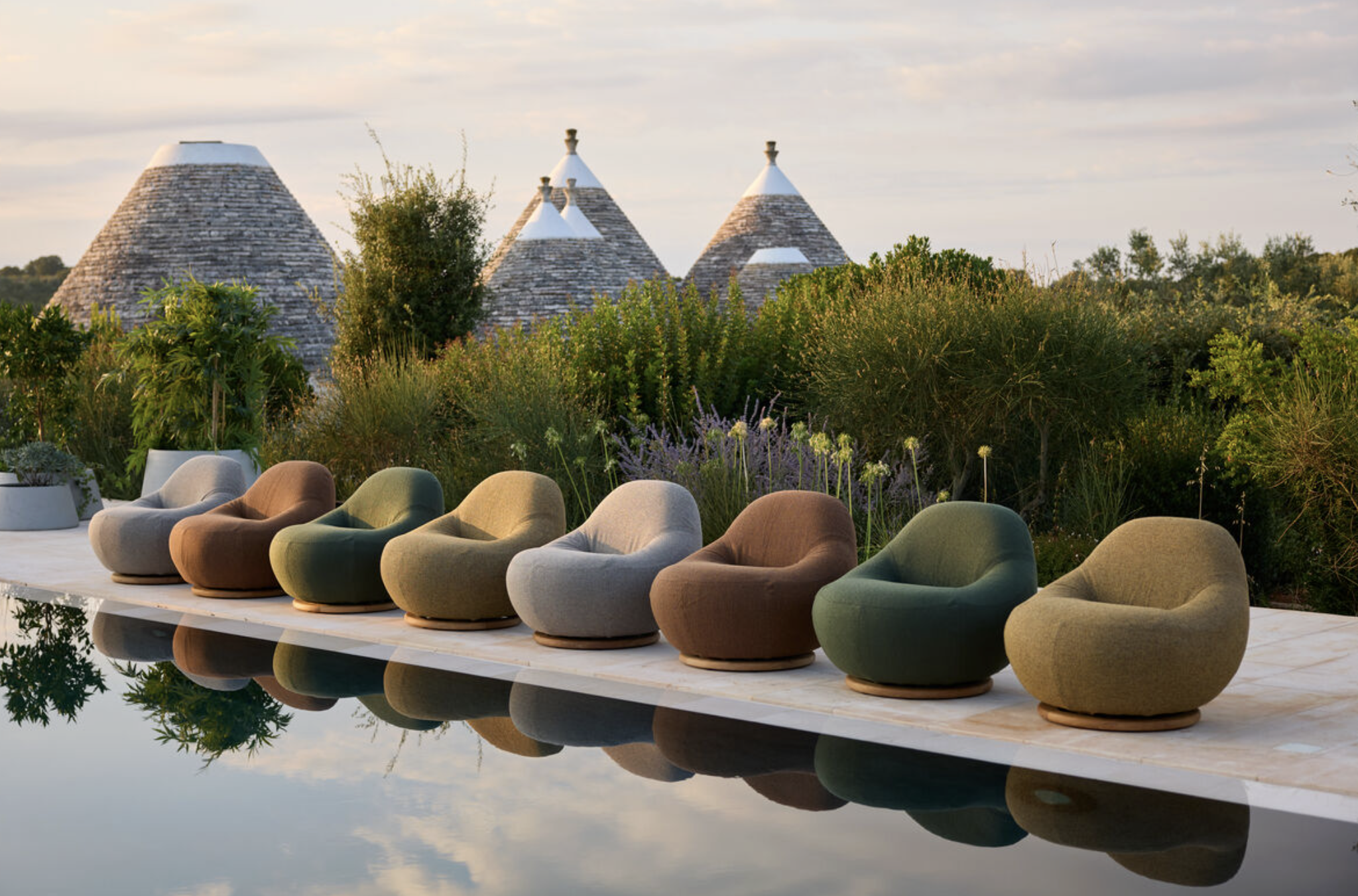 A scenic outdoor area features modern, rounded seating in various earth tones, lined along a reflective pool that mirrors the sky, surrounded by lush greenery and plants. In the background, there are traditional stone cone-shaped buildings with white roofs, set against a sky with soft clouds during the golden hour.