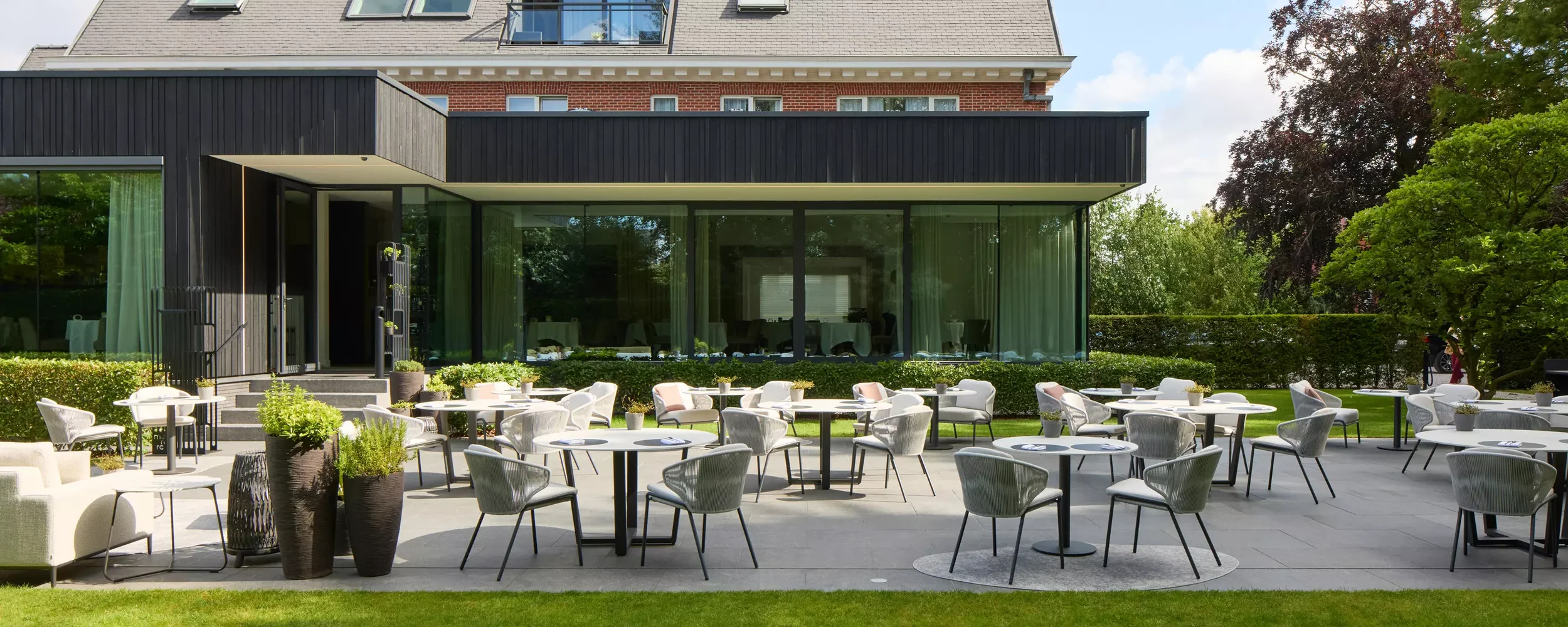 Outdoor patio with round tables and chairs, potted plants, and a modern building with large glass windows in the background