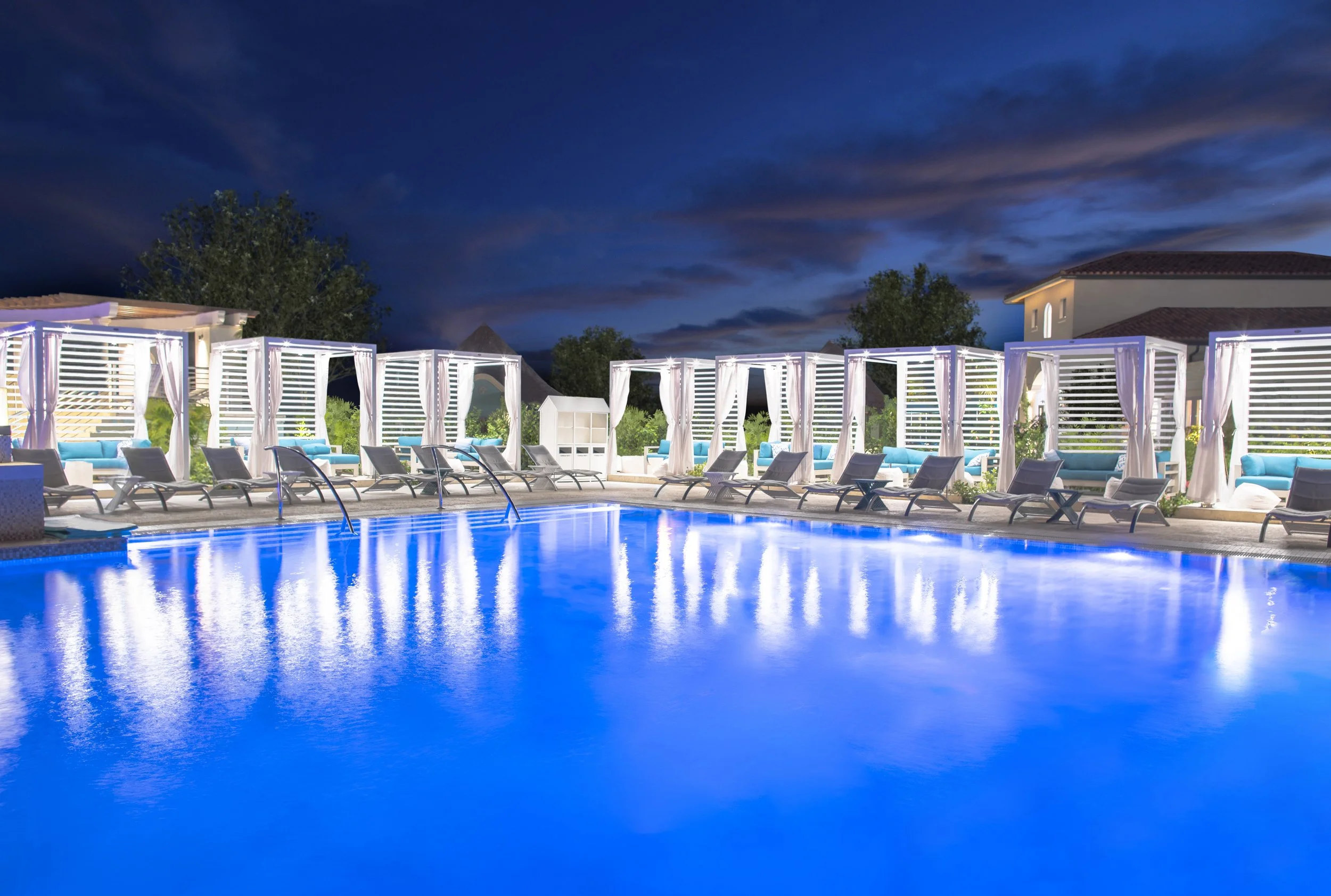 Pool area illuminated at night with lounge chairs and cabanas with curtains, surrounded by trees and residential buildings under a dark, cloudy sky.