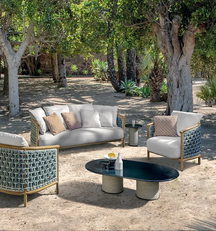 Outdoor living space with a modern sofa and chairs featuring woven sides, placed around a black oval coffee table and a small side table, under large leafy trees and desert plants in a sandy area.
