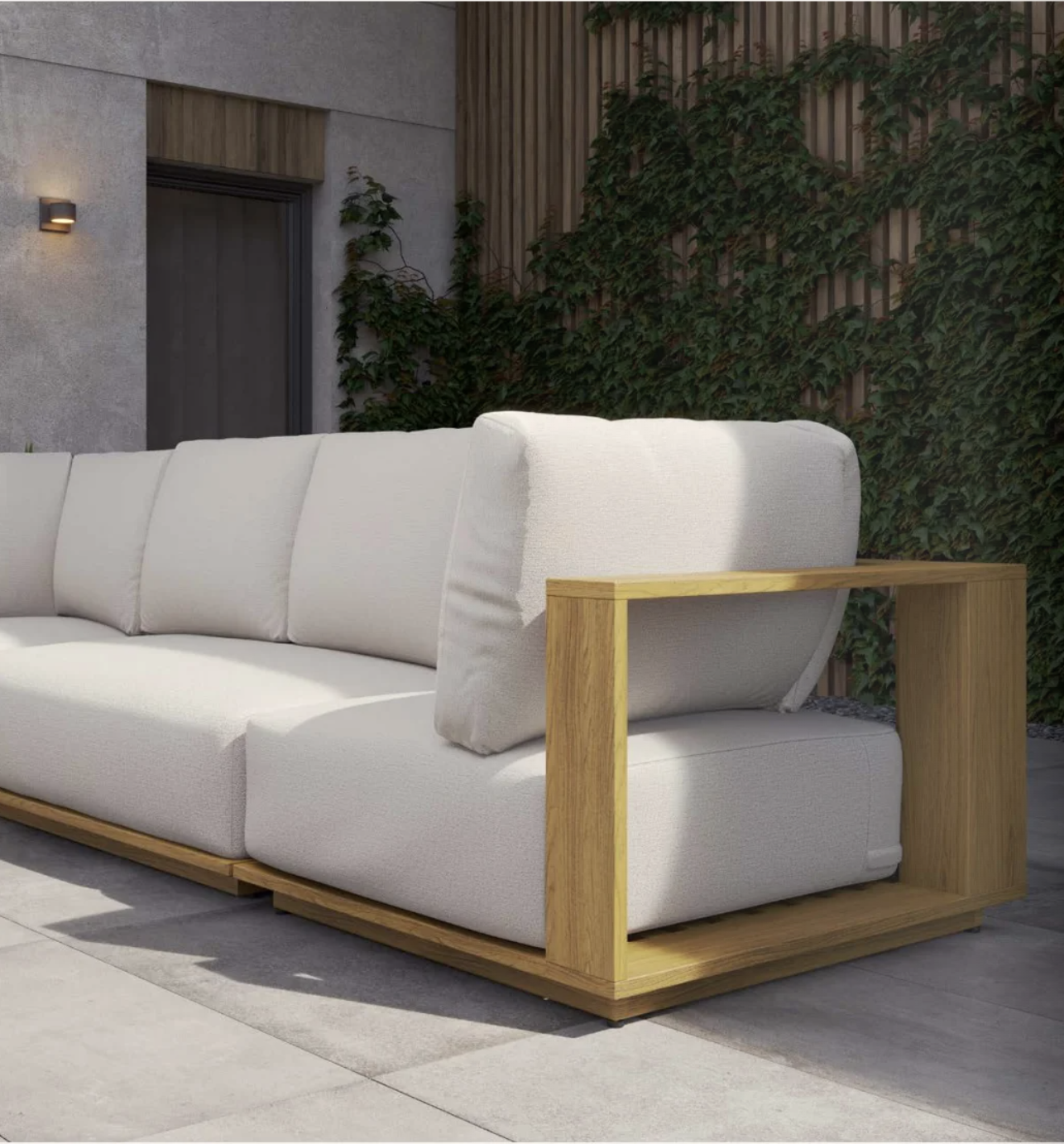 Outdoor patio sofa with beige cushions and wooden armrest, against a background of a wall with greenery and a door.