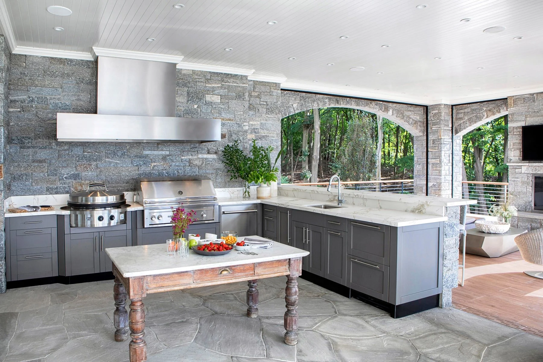 Modern outdoor kitchen with gray cabinets, white marble countertops, stainless steel appliances, and stone walls. There are large windows showing a wooded backyard, and a small wooden table with a fruit bowl, flowers, and food items on it.