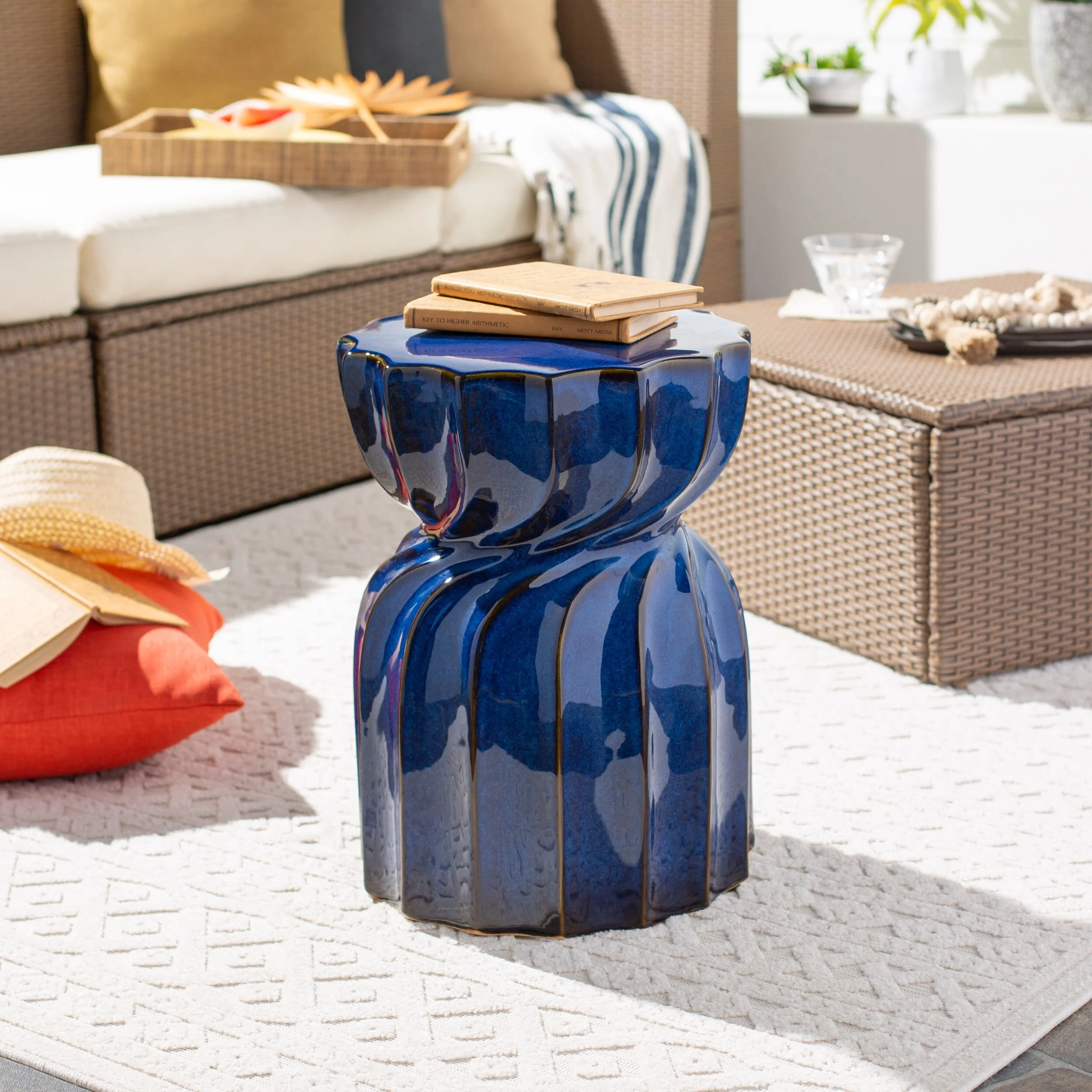 Close-up of a blue ceramic side table with a stack of three books on top, placed on a white textured rug in a cozy living space with a sofa, pillows, a tray with decorative items, and plants in the background.